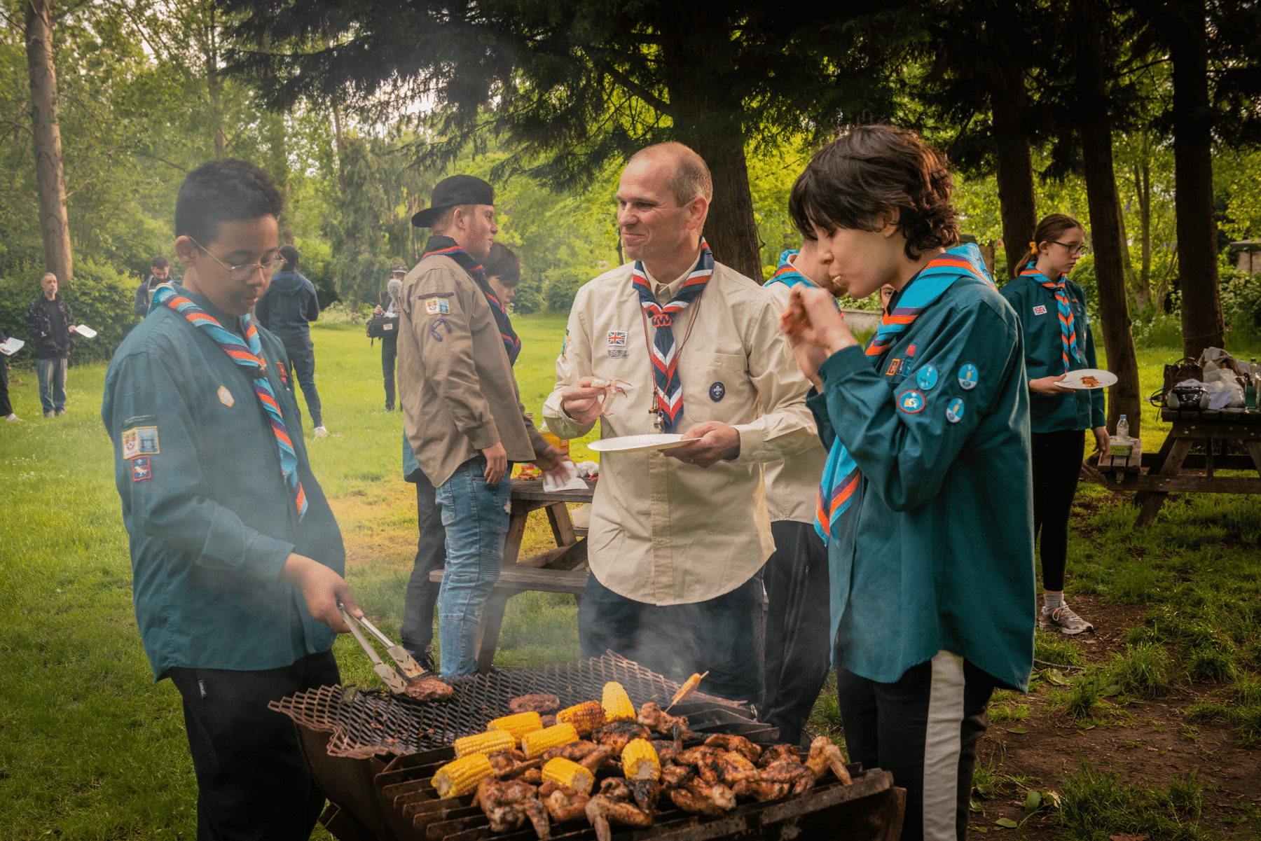 Two Scouts and their leader are at a big Scout event. They are stood around a BBQ eating, with the leader in the middle. There is lots cooking on the BBQ, including sausages and corn on the cob. One Scout is moving food around using tongs and the leader is smiling at him, looking very proud.