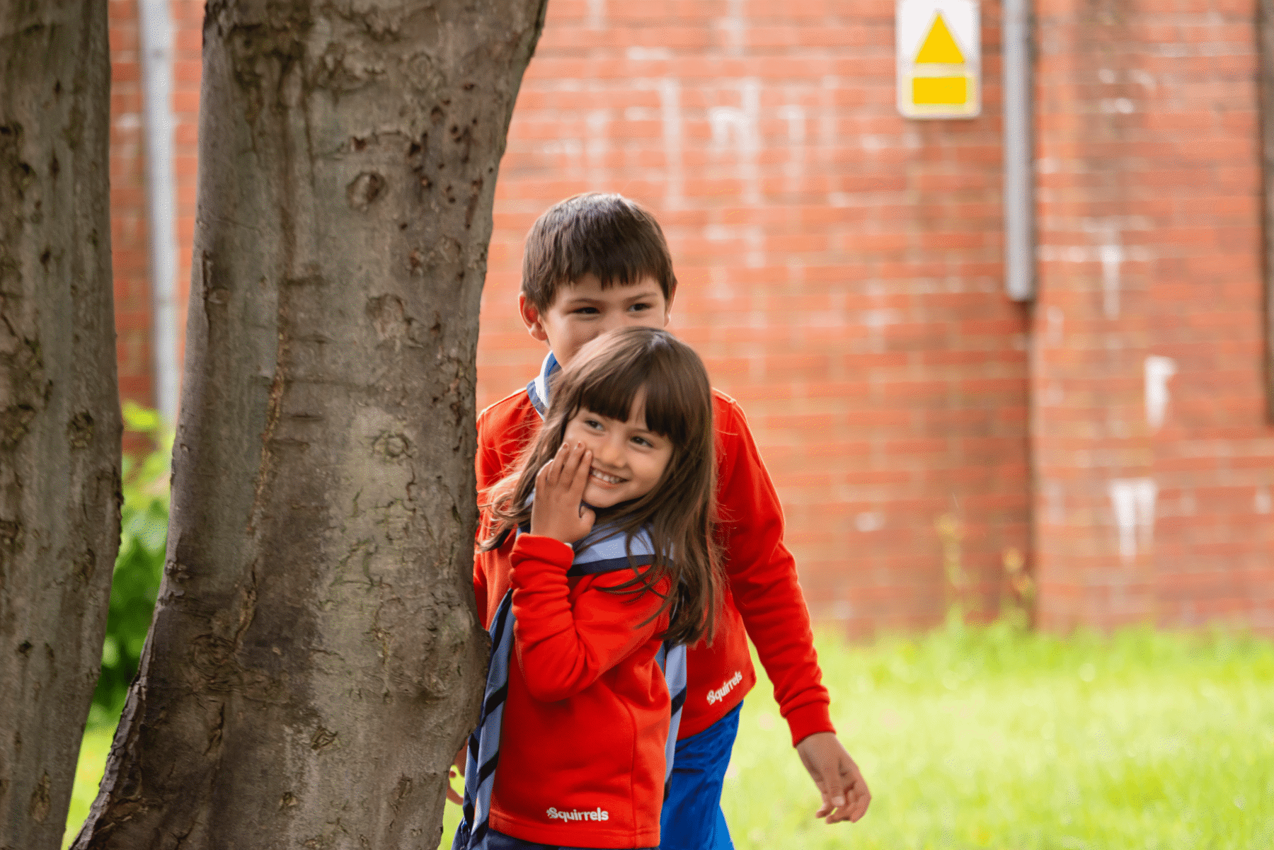 Two Squirrels, a boy and a girl, are hiding behind a tree and leaning out so that we can see their faces. They are playing a game and have big smiles on their faces. They are wearing red jumpers and the girl has her hand over part of her mouth.