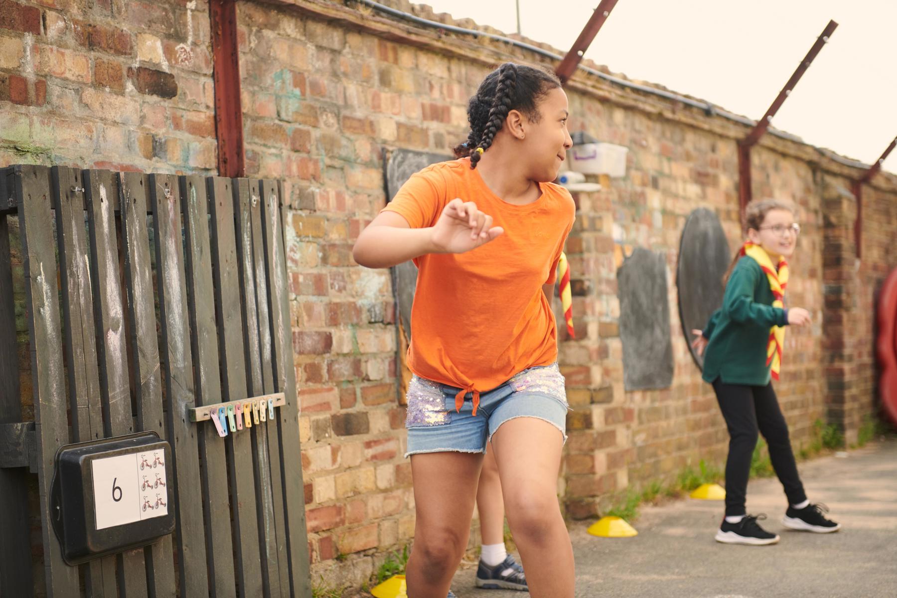 A Cub group is playing a running game outside their meeting place on a playground. There are two girl Cubs playing the game. The girl in the foreground is wearing an orange t-shirt and is looking over her shoulder as she runs. The girl in the background is wearing her green Cub top and looks ready to start running.