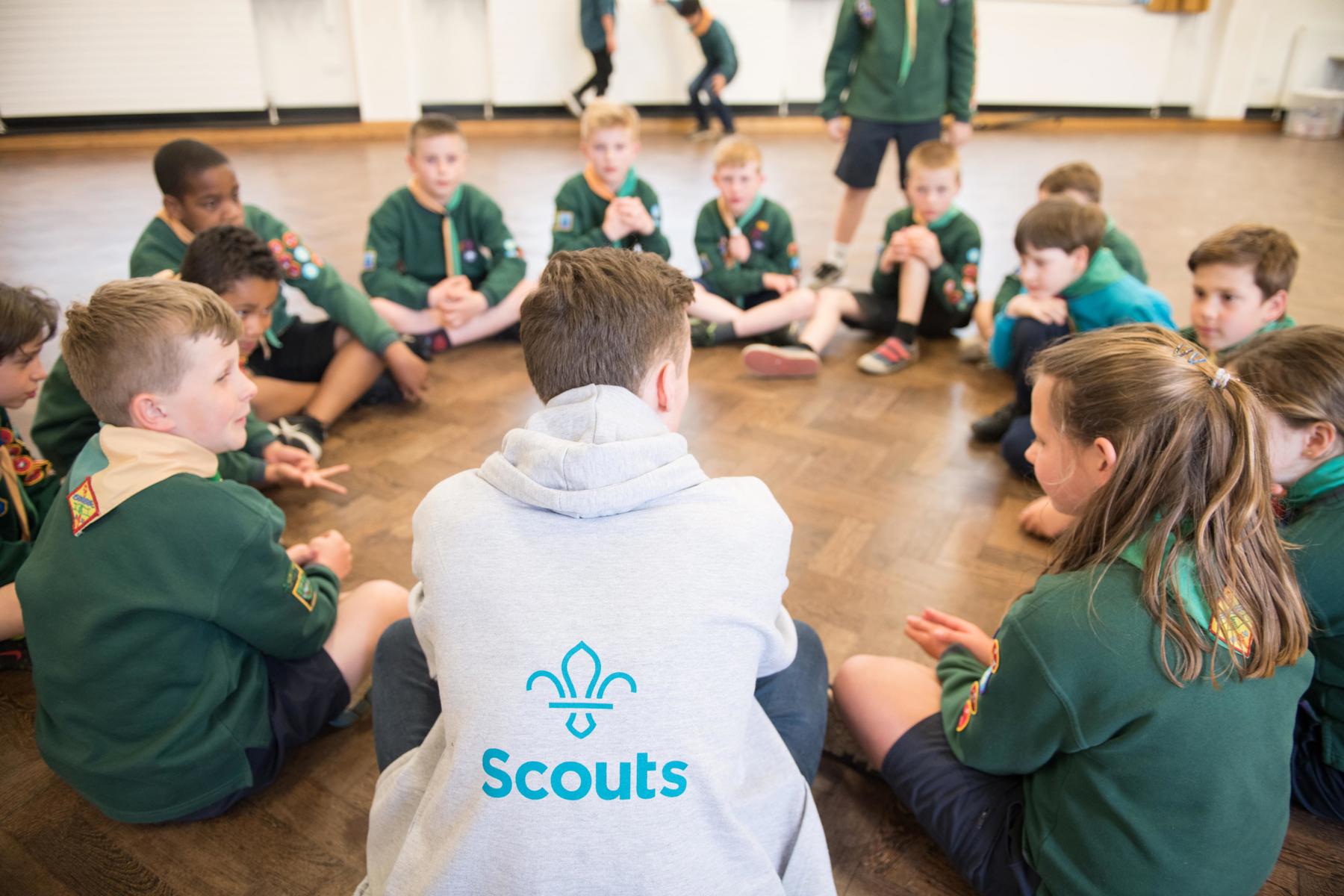 A group of cubs are sat in a circle on the wooden floor of their meeting place. Their male leader is sat in the circle, with his back to the camera. We can see the Scouts logo on the back of his hoodie. They are all sat in the circle talking.