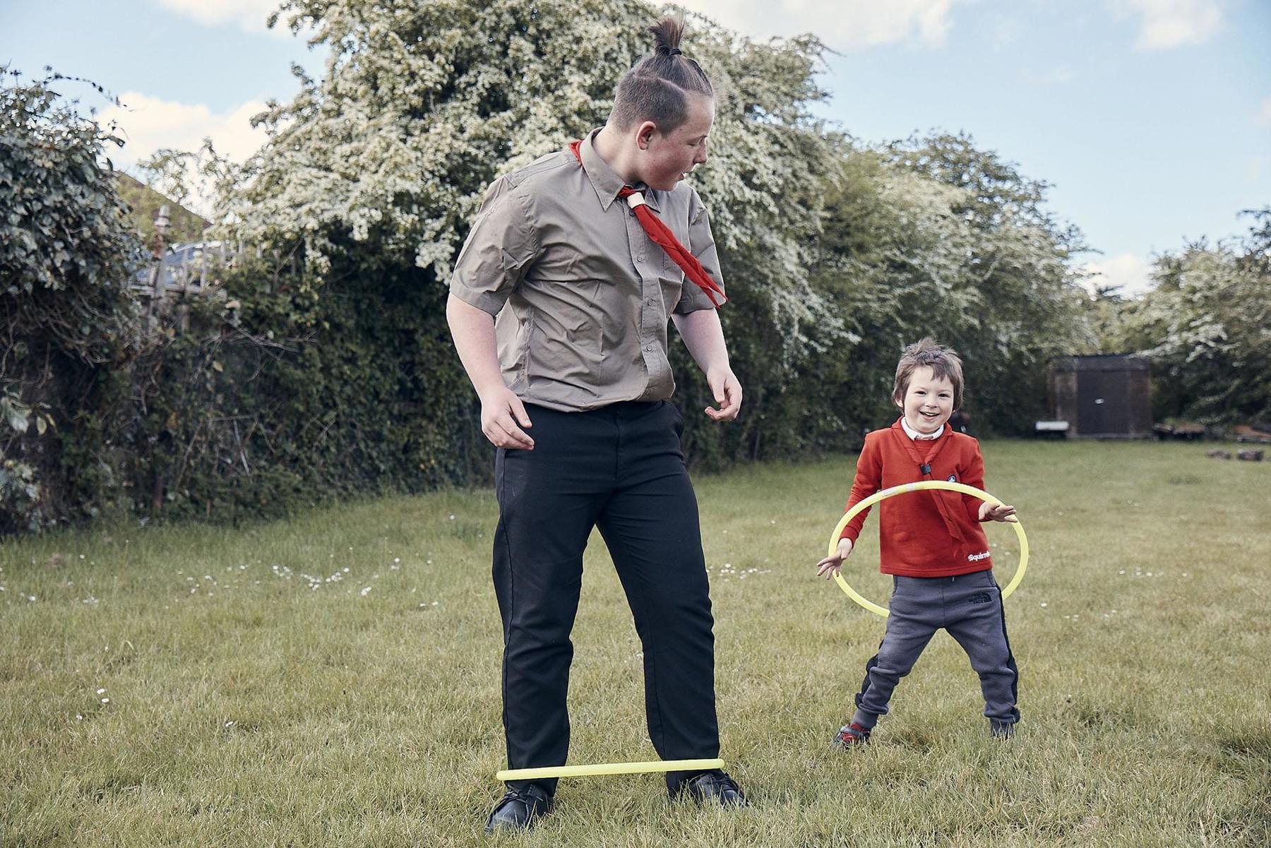 A Scout and Squirrel are stood on a field in the grass, and they are trying to hoola hoop. The Scout has the hoop around his ankles and the Squirrel is trying to hoola hoop around his waist. They are both smiling and having fun.
