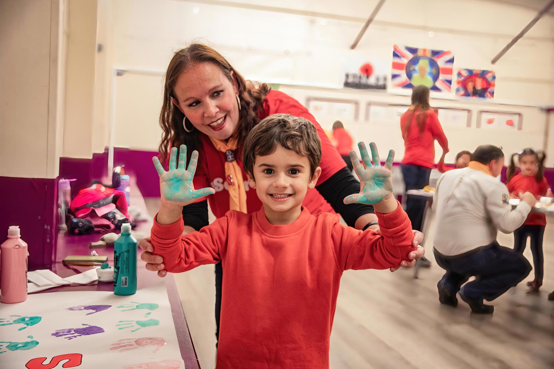 A boy Squirrel is looking at the camera smiling, with both hands held up in the air. His hands are covered in green paint and he has been making handprint pictures. A female volunteer is stood behind him, smiling. They are inside their meeting place.