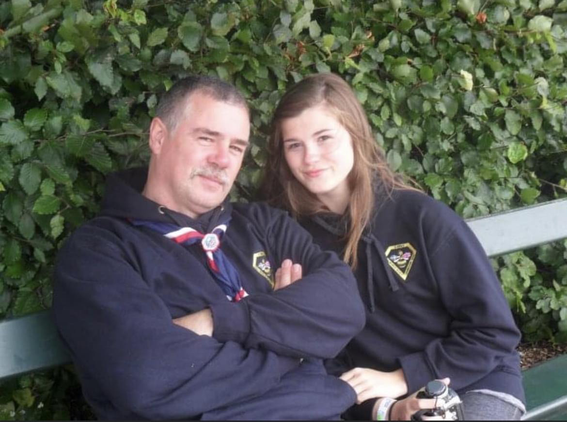 Steve and his daughter are sat on a green bench together. They are both wearing Scout hoodies and Steve is wearing a blue, red and white necker. They are both smiling at the camera and are outside in front of a tall bush.