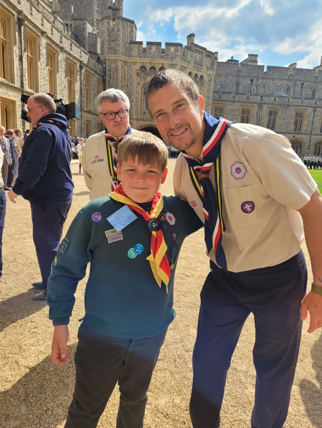 Bear Grylls and Jack Lally put an arm round each other, both wearing Scout uniforms, in front of Windsor castle