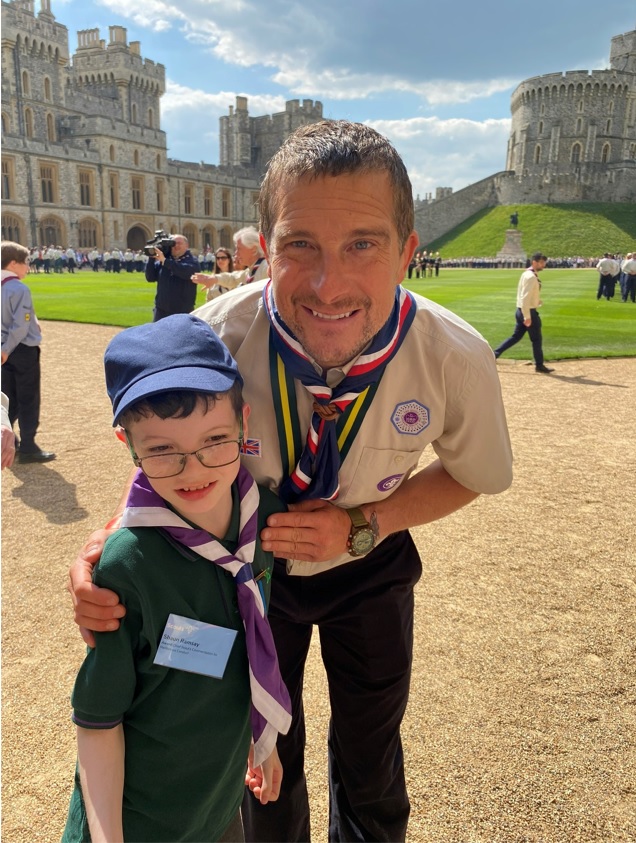 Bear Grylls puts an arm round Shaun Ramsay, both wearing Scout uniforms and neckers, in front of Windsor castle