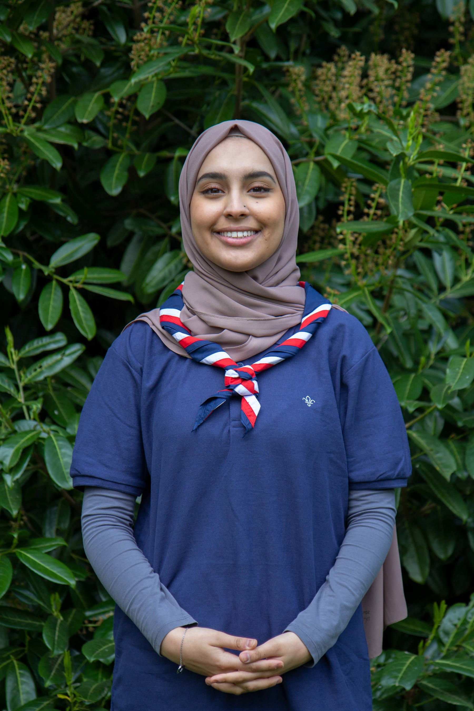 Ayesha wearing a Scouts polo and necker, standing in front of bushes, smiling at the camera