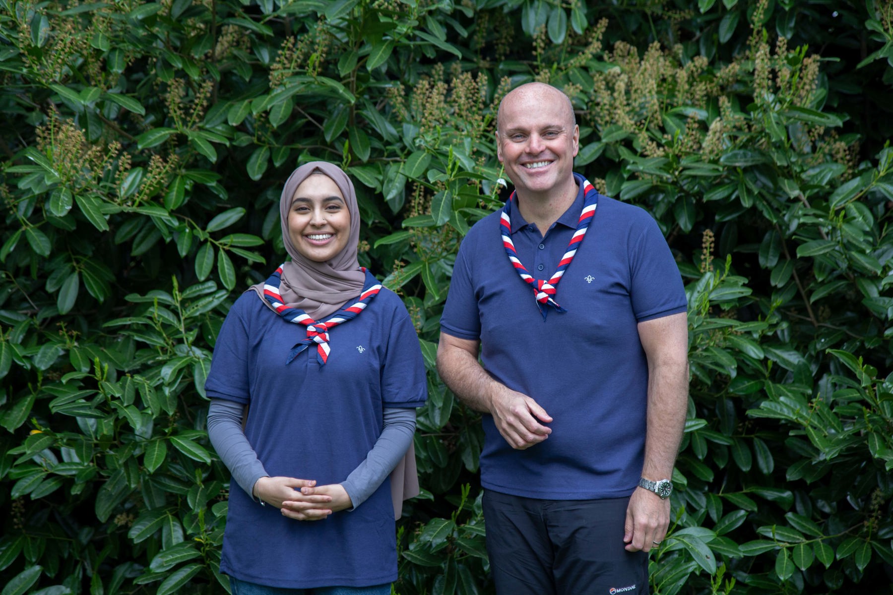 Ayesha and Carl wearing Scouts polo shirts and neckers, standing in front of bushes, smiling at the camera