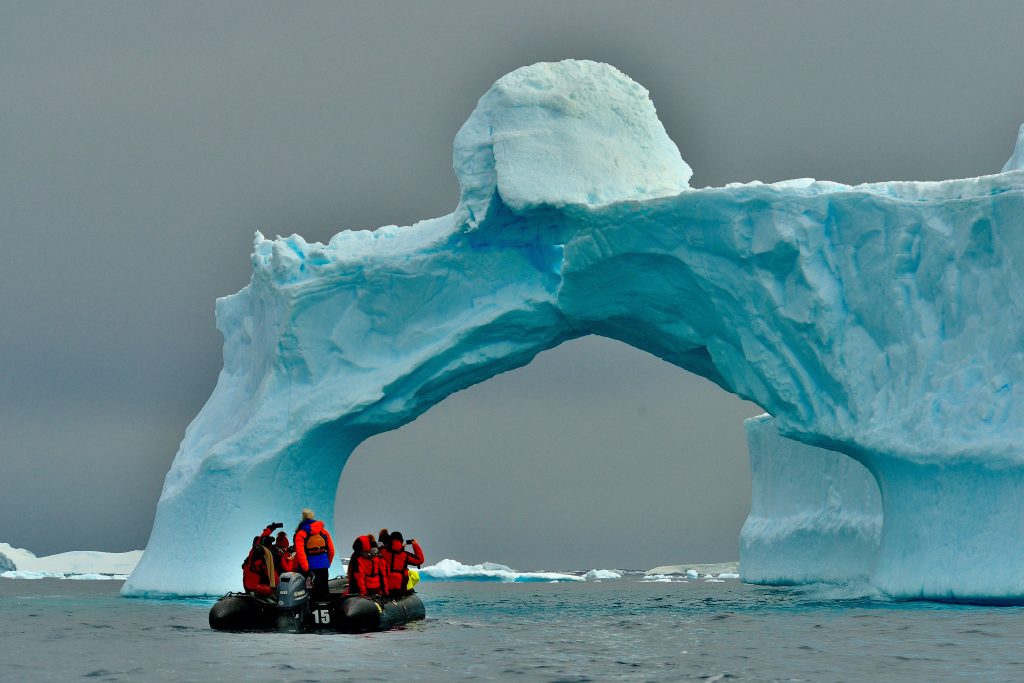 Polar explorers in a small boat on the water beneath an ice arch in Antarctica