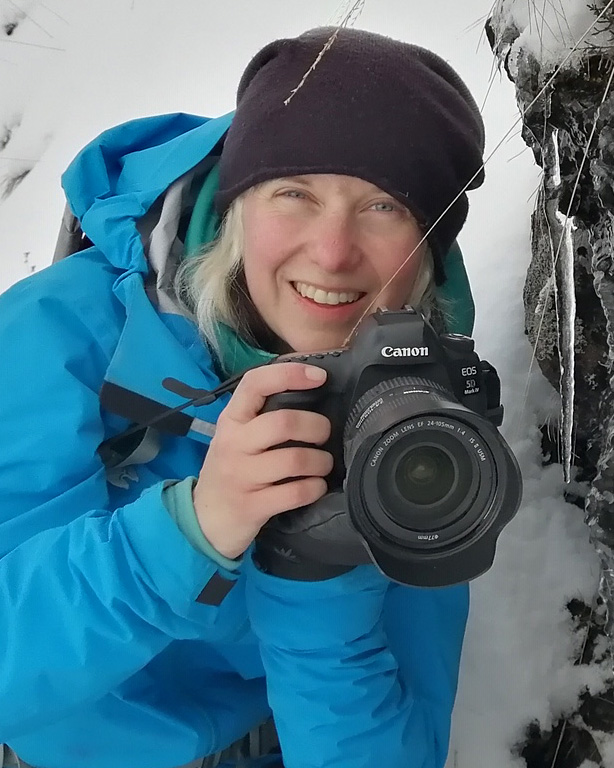 Phoebe Smith smiles pointing a Canon camera towards the camera, with an icy background behind, and wearing clothes for cold weather 