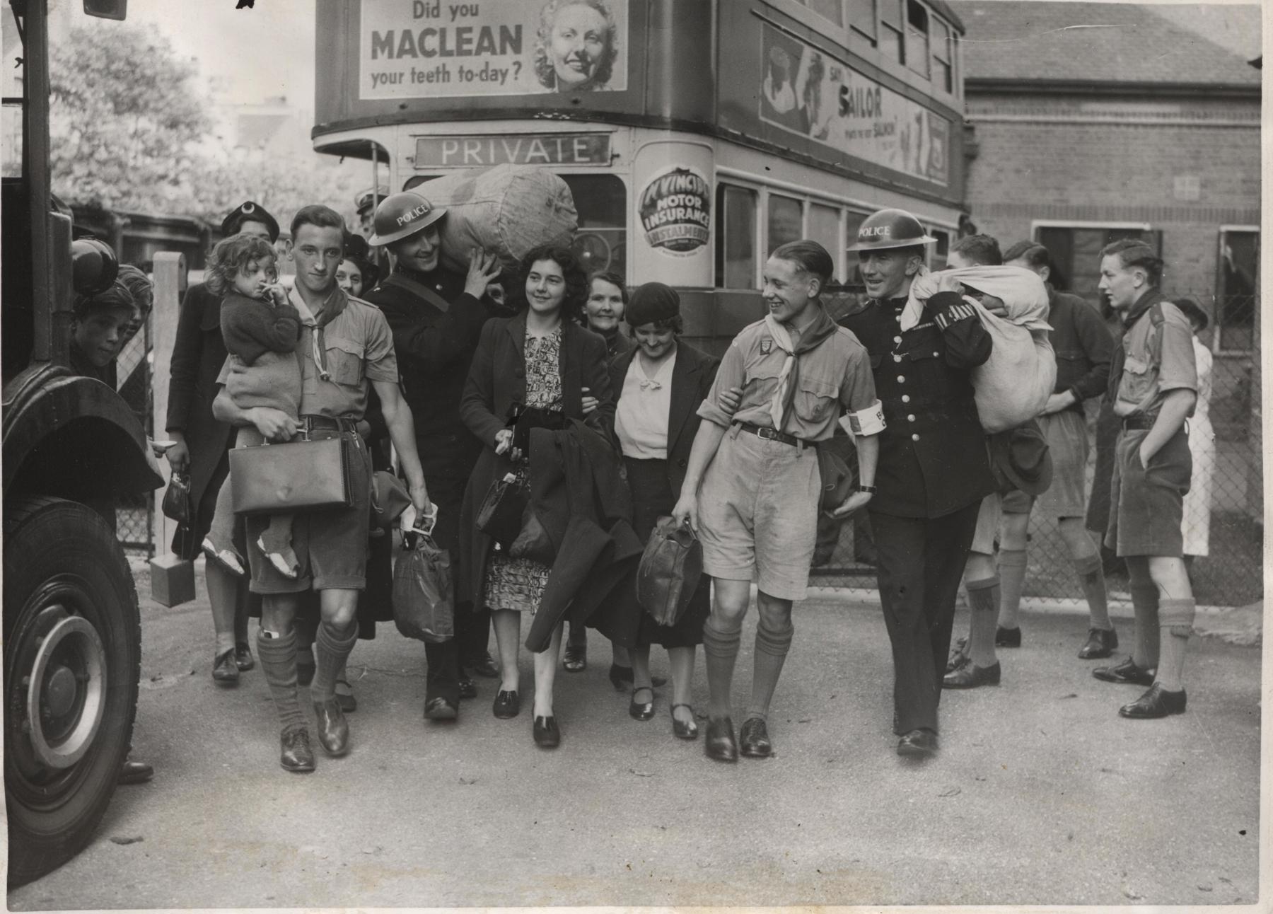 Scouts help to carry the luggage of Second World War evacuees in front of a bus. The Scouts are in uniform and they are walking towards the camera with children and women. One is carrying a suitcase and another is carrying a young girl.