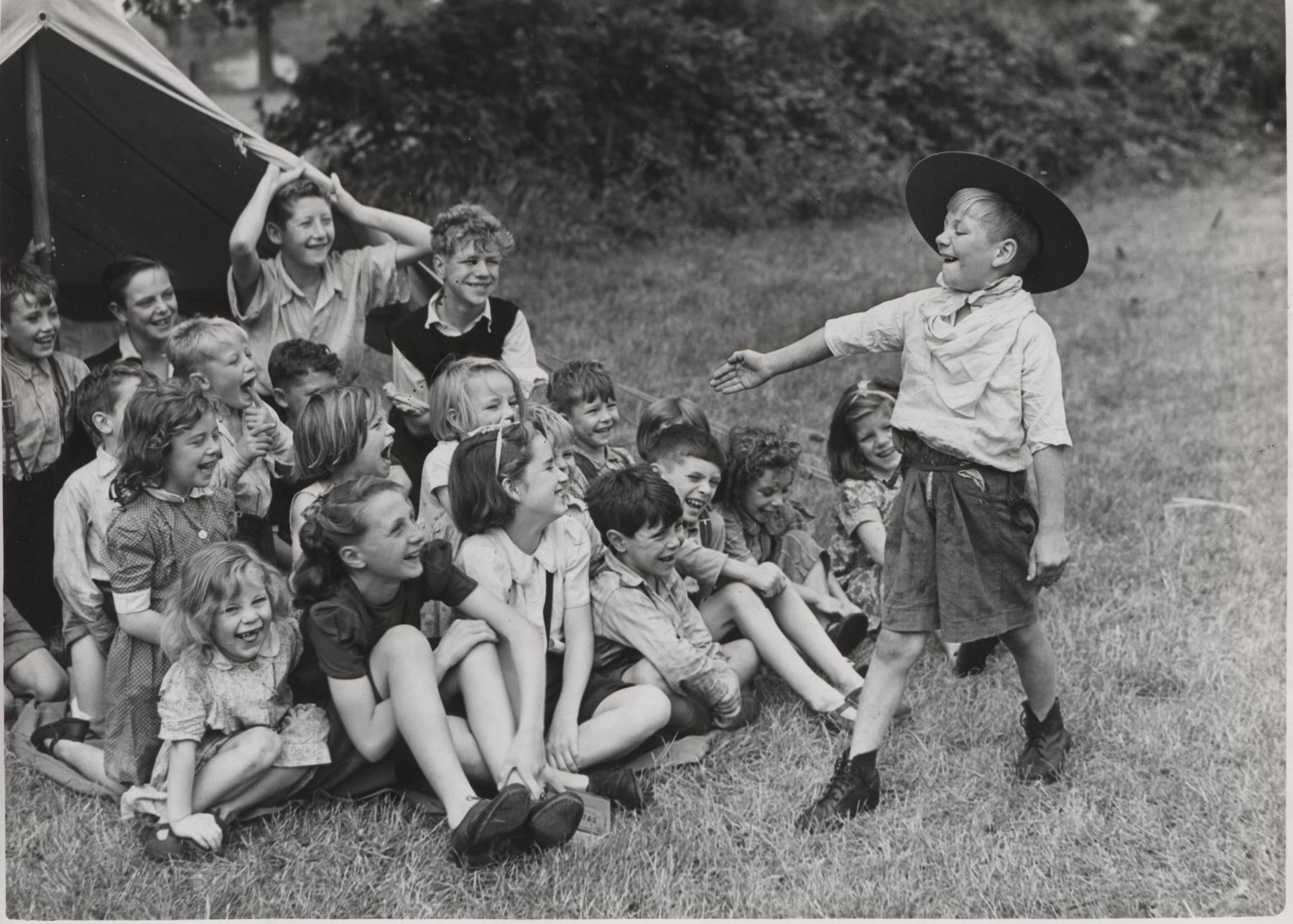 A group of young children sit on the grass in front of the tent. They are smiling and laughing as a young Scout boy is stood in front of them, playing and making them laugh.