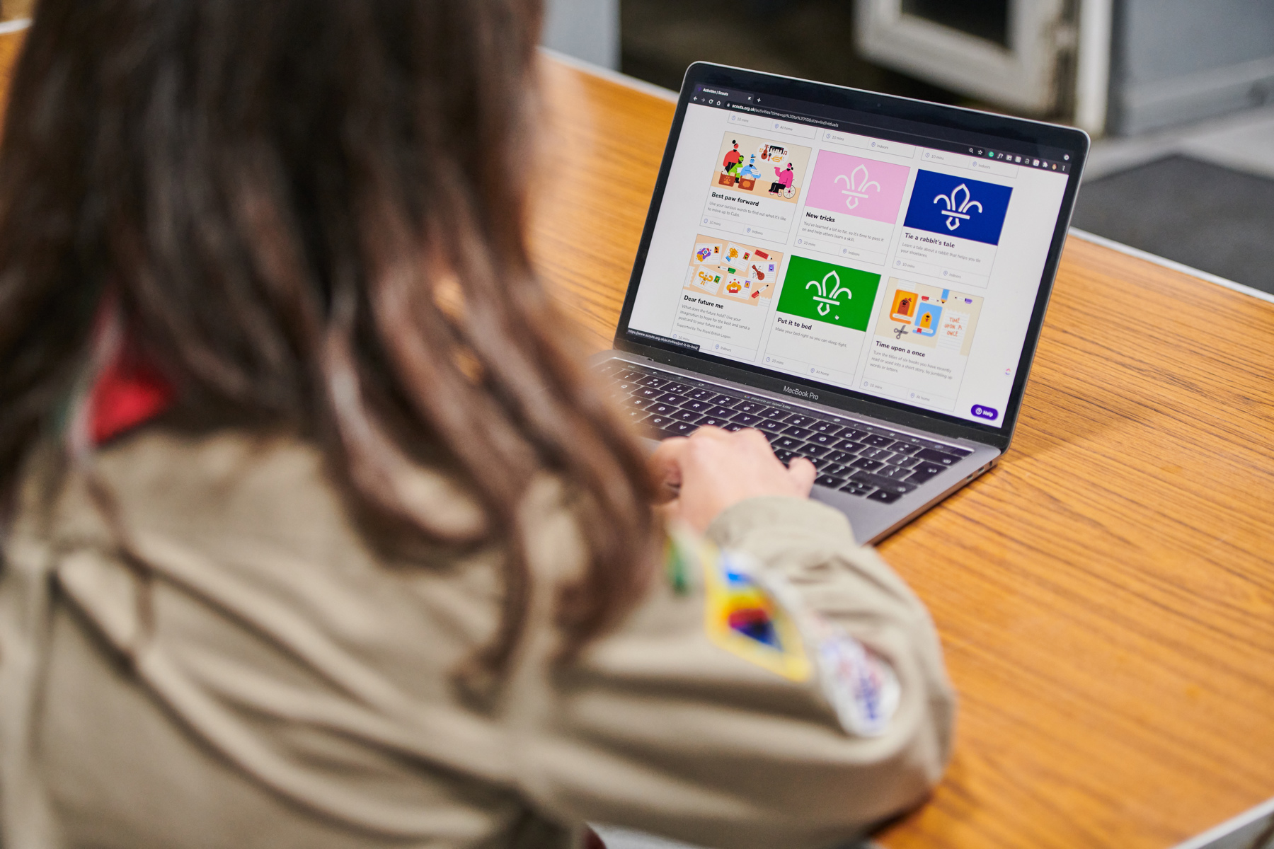 We can see the Scout activity finder website open on a laptop screen, with the laptop on a table. We're looking over the shoulder of a female Scout, who is scrolling through the page with her hand on the laptop.
