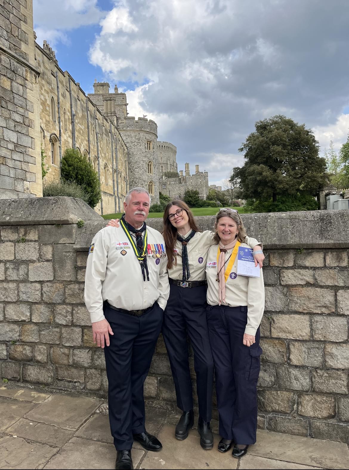 Hannah, Steve and Sue Ralph stood by one of the walls of Windsor castle. Hannah is in the middle with her arms round Steve and Sue, with one hand holding her award. They're all dressed in formal Scout uniform, with neckers, lots of badges and medals.
