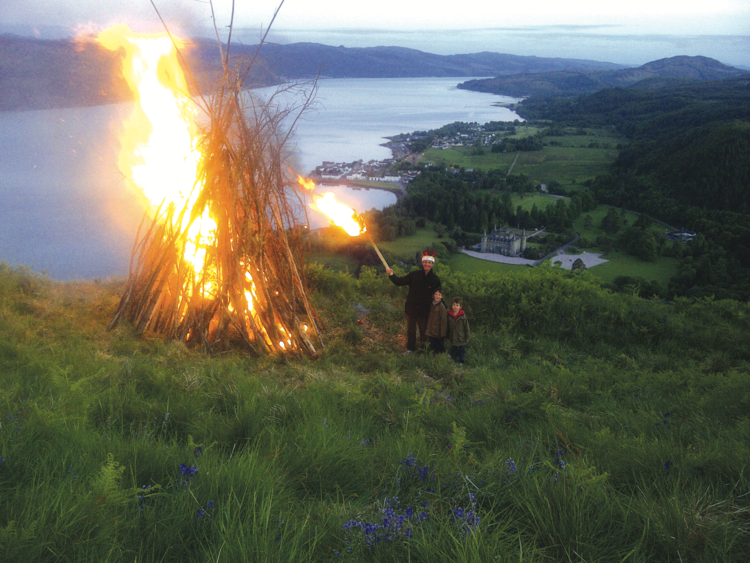 A large bonfire beacon aglow on a hill,  with an adult and two children next to it looking towards the camera, against a background of water and a castle below