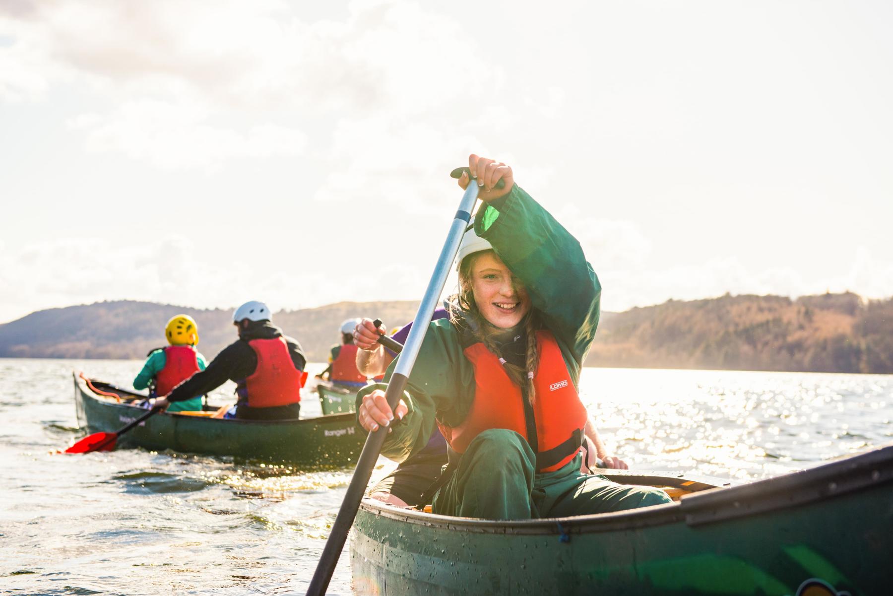 A group of Scouts are out on a lake in canoes. There are hills in the background and a cloudy sky. A young girl is in her canoe and facing the camera. She is smiling while pushing her oar into the water. She's wearing a life jacket and helmet and looks like she's having a lot of fun.