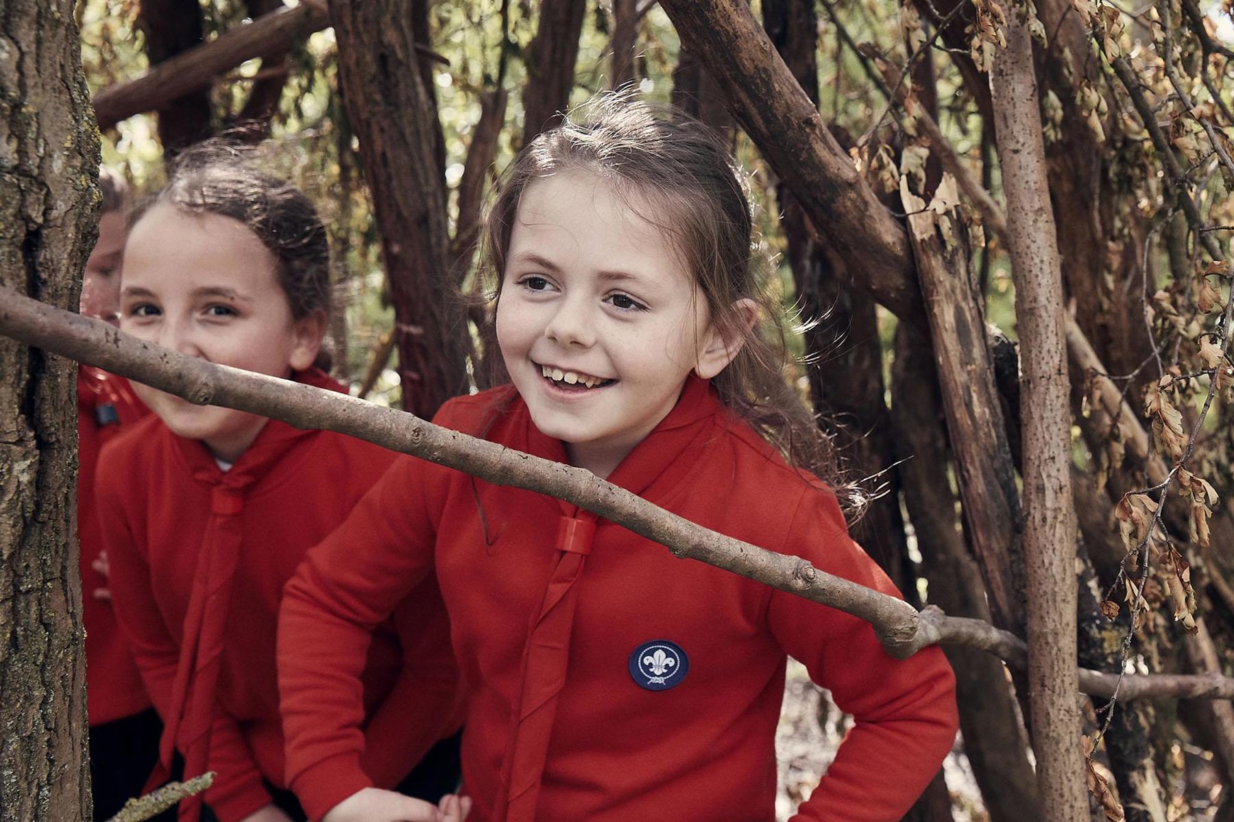 Two girls, who are in Squirrel uniform, are sat in their outdoor den and smiling at the camera. They're surrounded by branches, leaves and tress while sat inside the den.