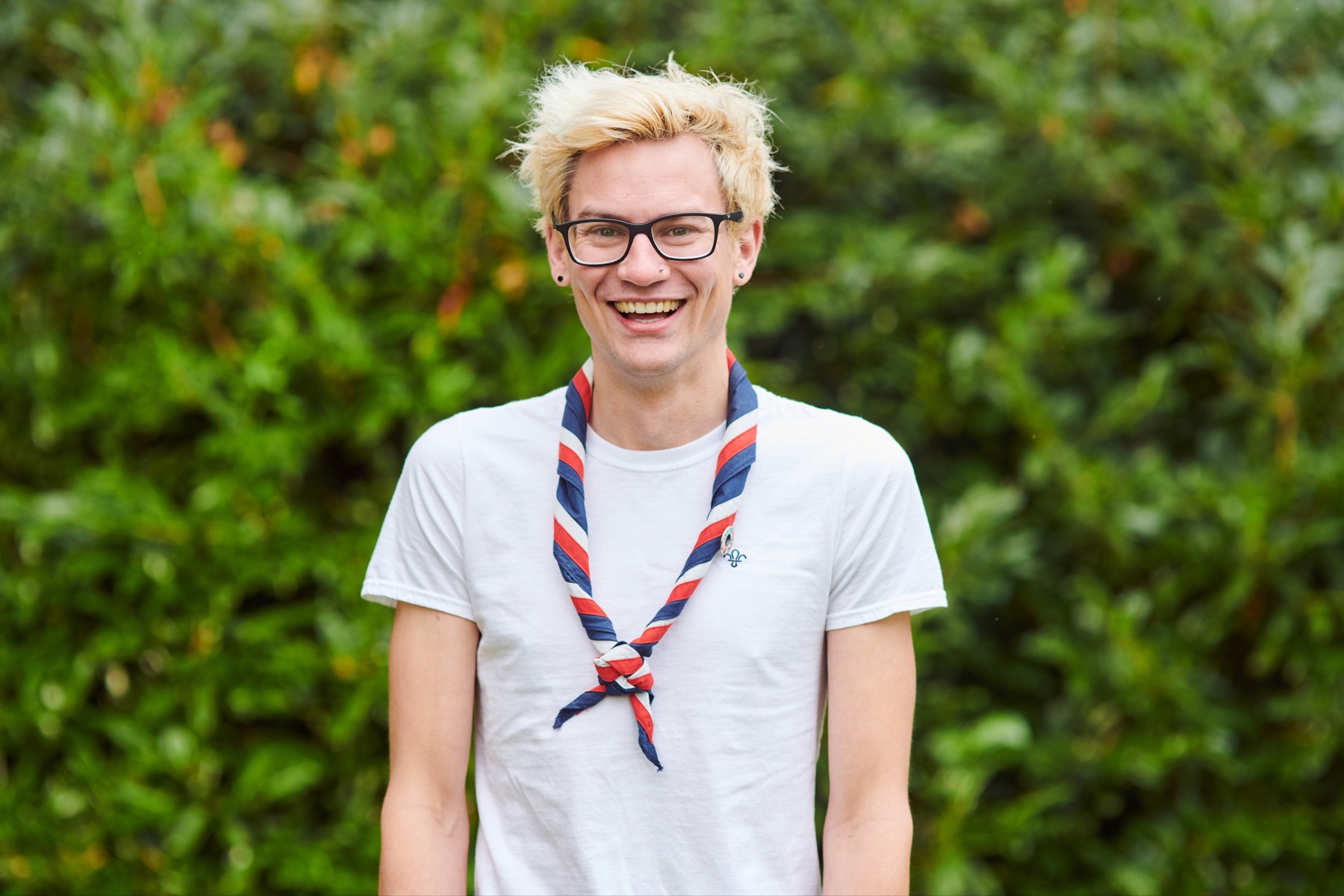 A Scouts volunteer outside wearing a white t-shirt and necker, smiling in front of bushes
