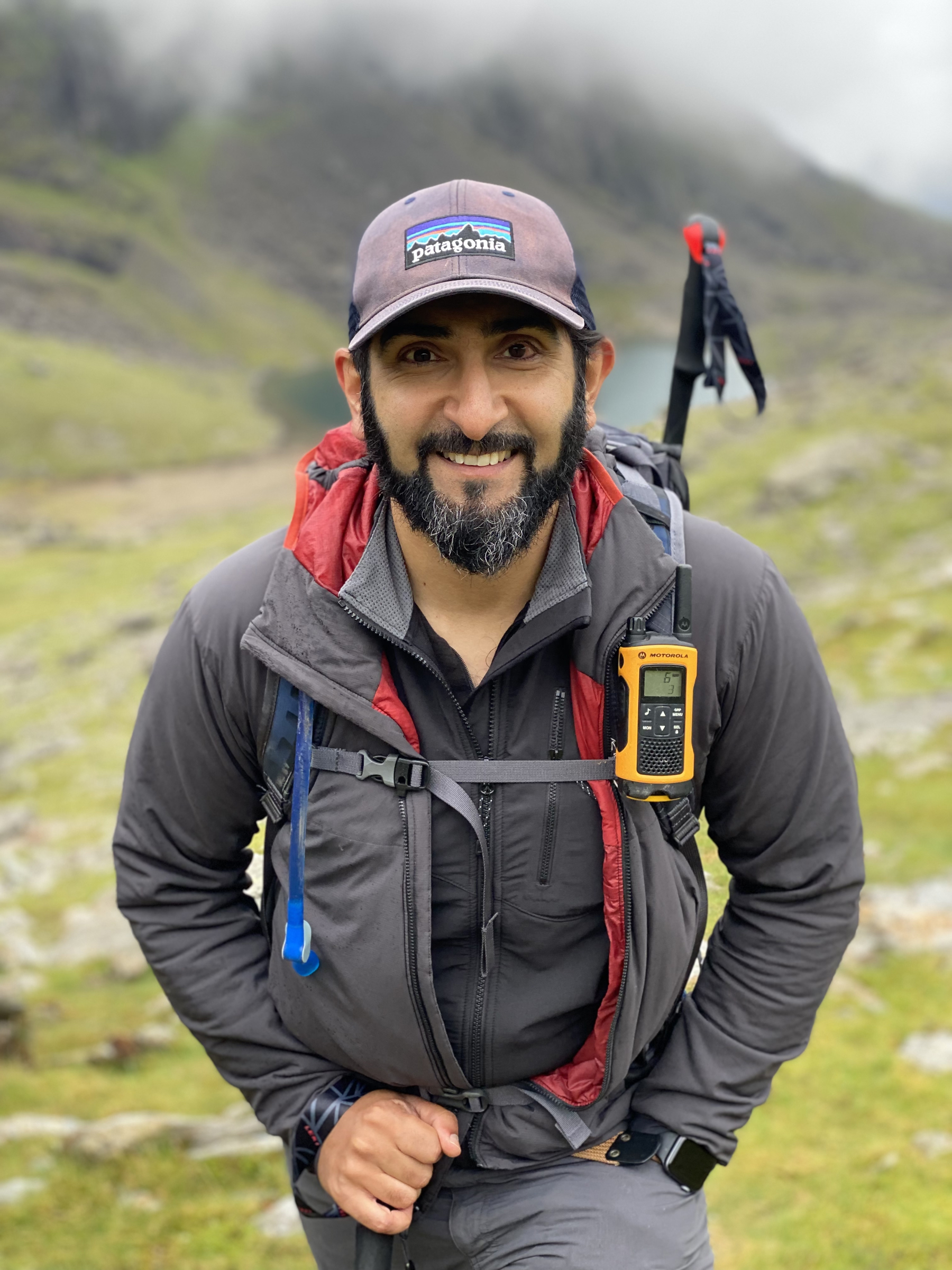 Mahroof is in his hiking gear, with GPS, hat, walking poles and waterproofs. He’s stood facing the camera, with cloud covered mountains and a small lake behind him.