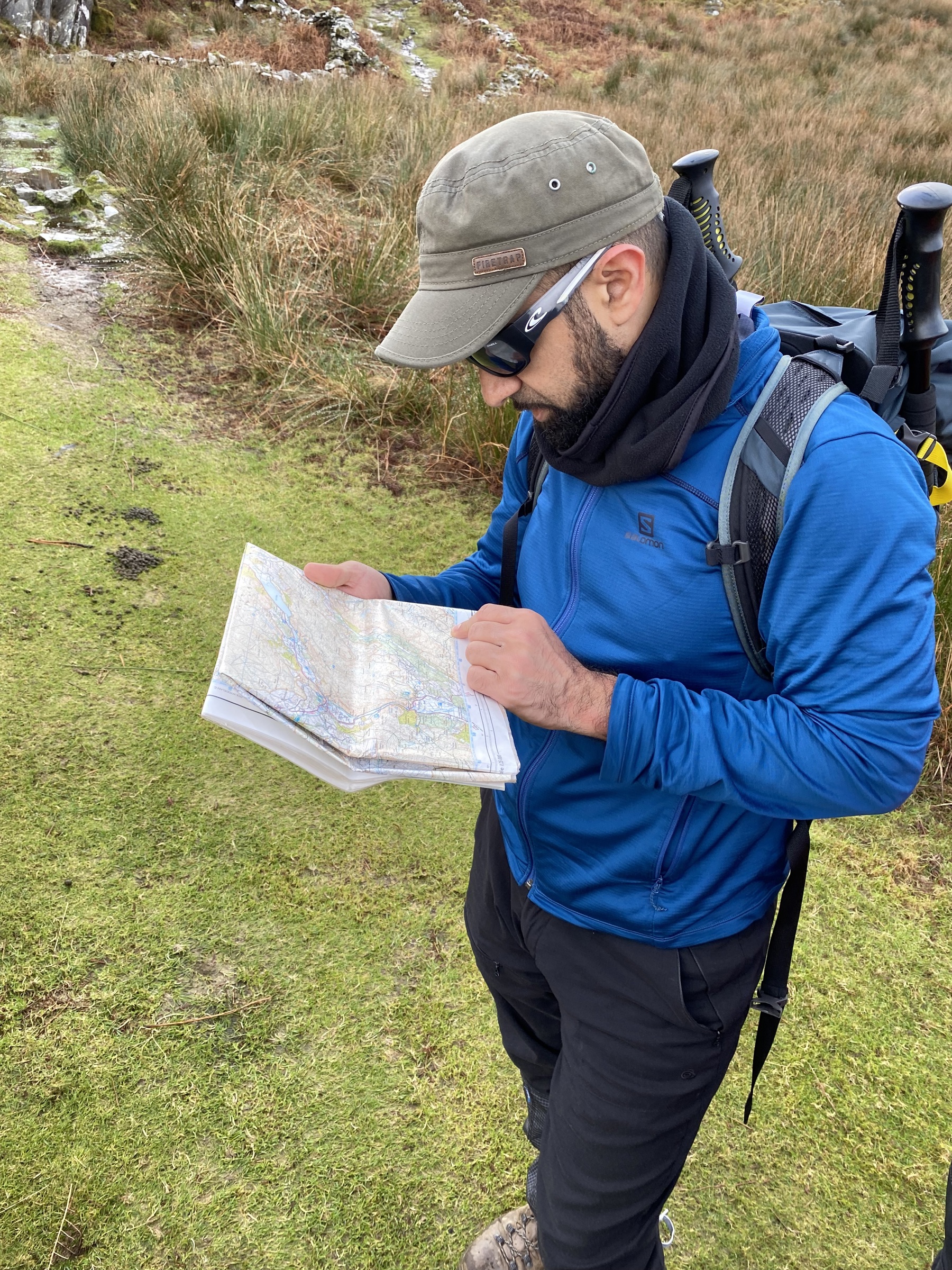 Mahroof is out in a field, with some marshland behind him. He’s looking at the map, while wearing his walking gear, boots, hat and sunglasses. He’s wearing a blue waterproof and his walking poles are in his bag.