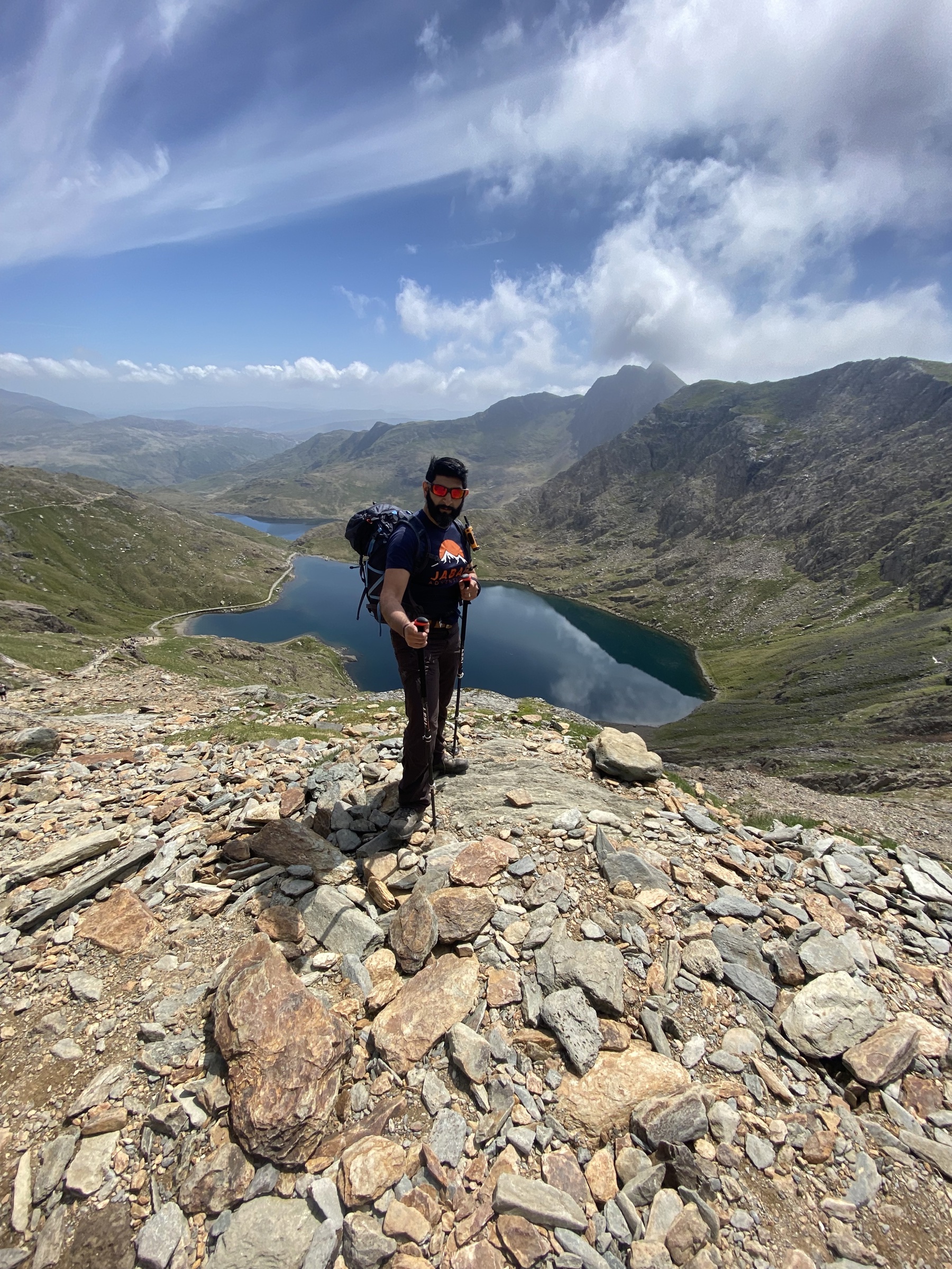 Mahroof is stood on a rock pile on top of a mountain. There’re mountains surrounding a lake behind him, beneath a blue sky. Mahroof has his walking poles, backpack and sunglasses.