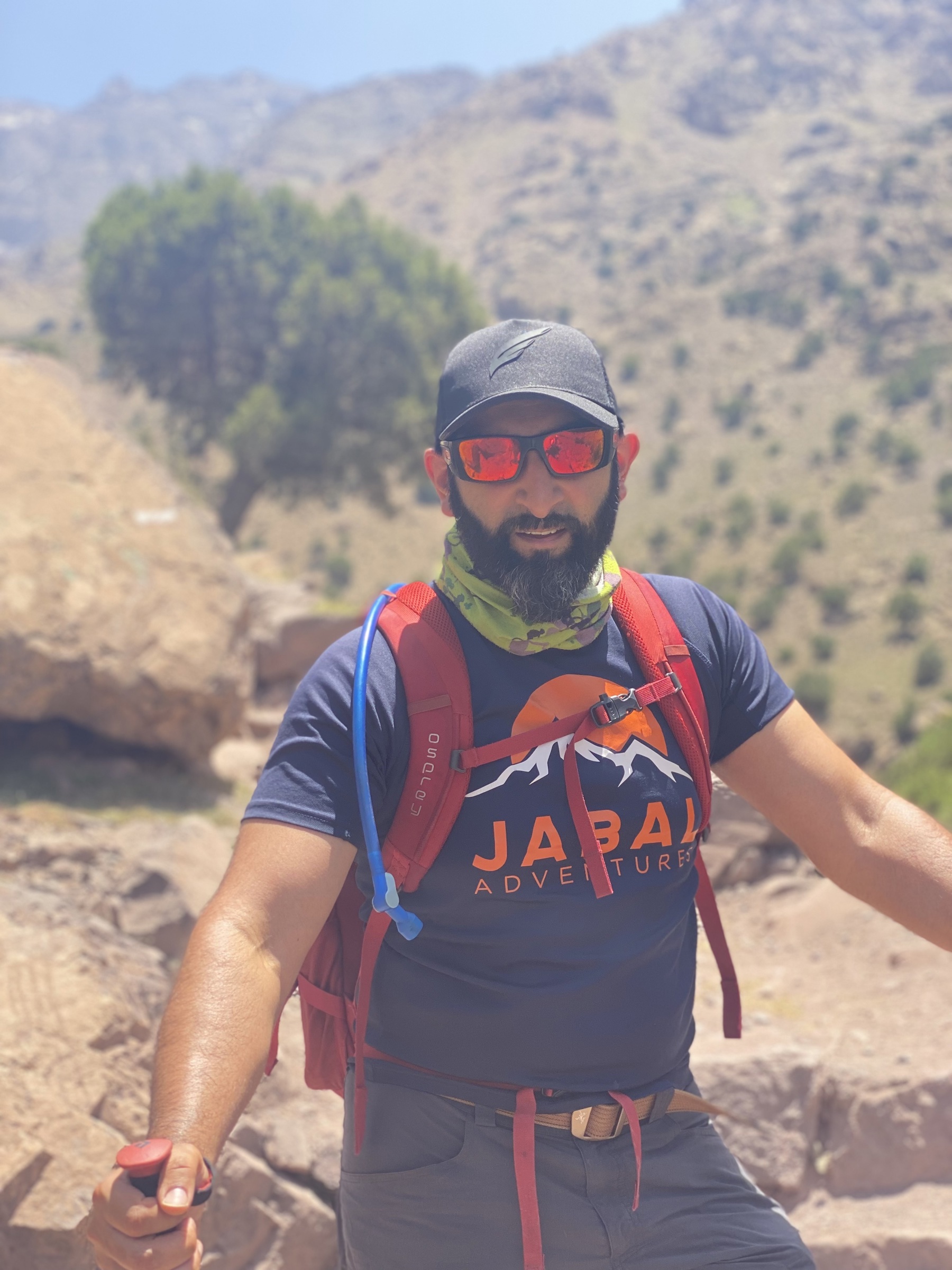 Mahroof is hiking in hot weather. He’s wearing sunglasses and a hat, while stood in front of sandy rocks and sparse trees.