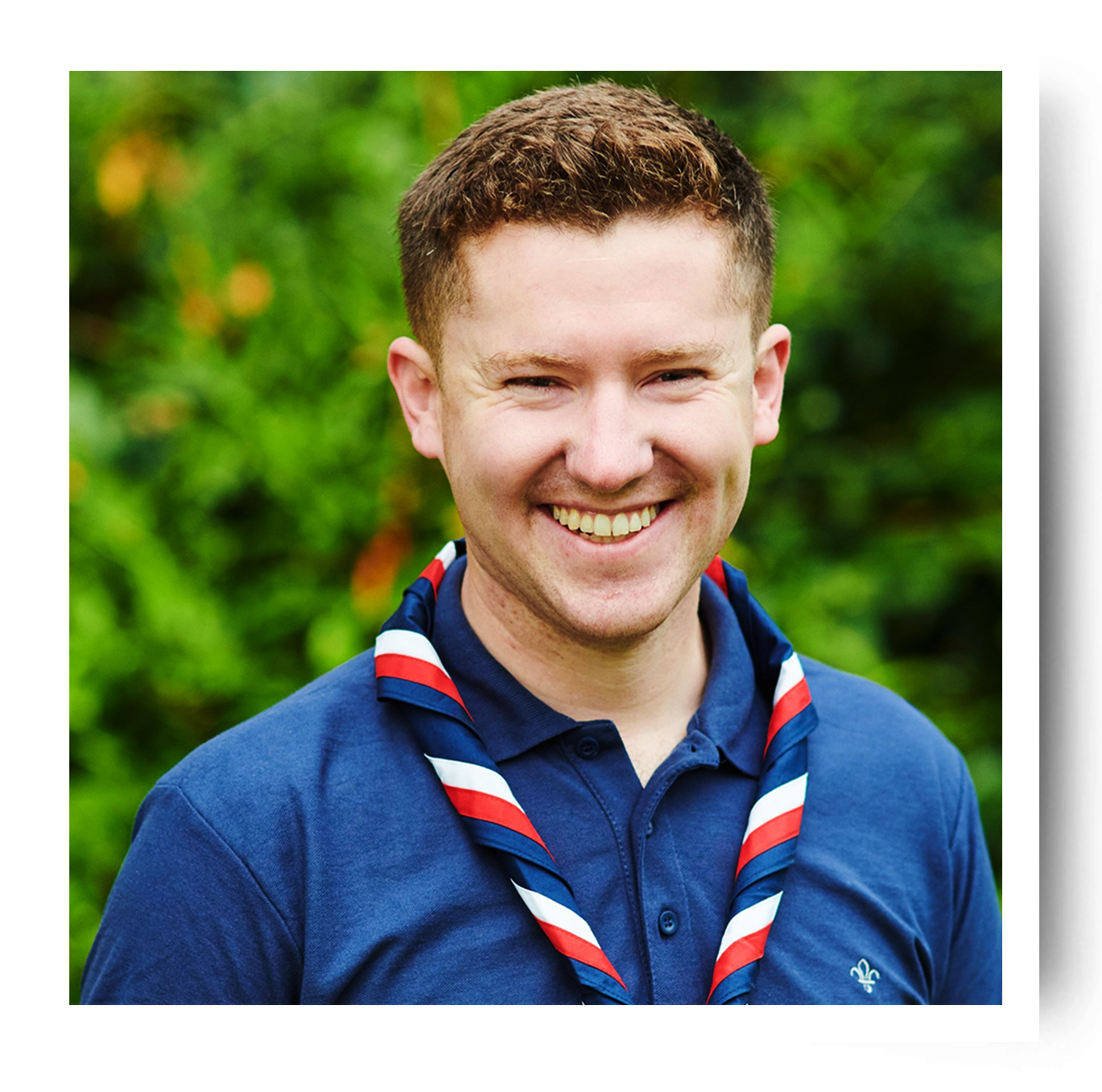 UK Youth Commissioner Ollie Wood is outside wearing Scouts uniform in front of a hedge and is smiling at the camera