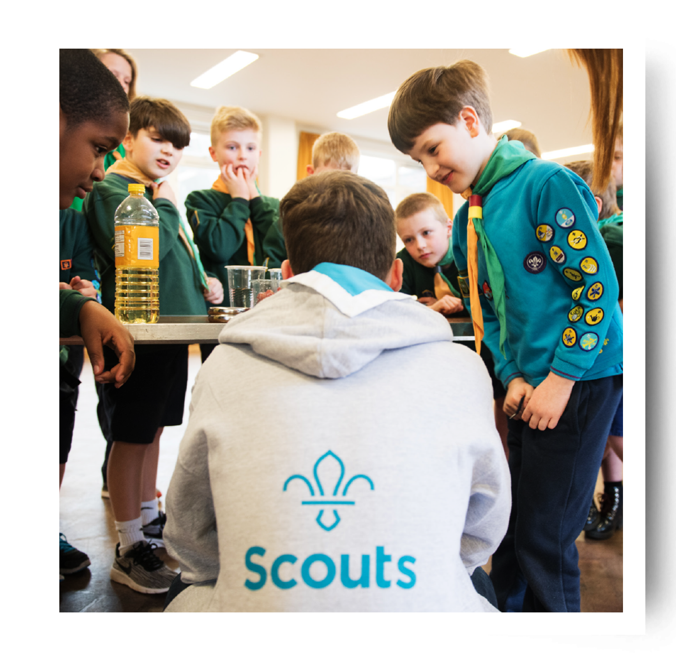 A volunteer is crouching with his back to the camera wearing a grey hoodie with the Scout logo. Beavers are standing around him wearing their uniform and badges.