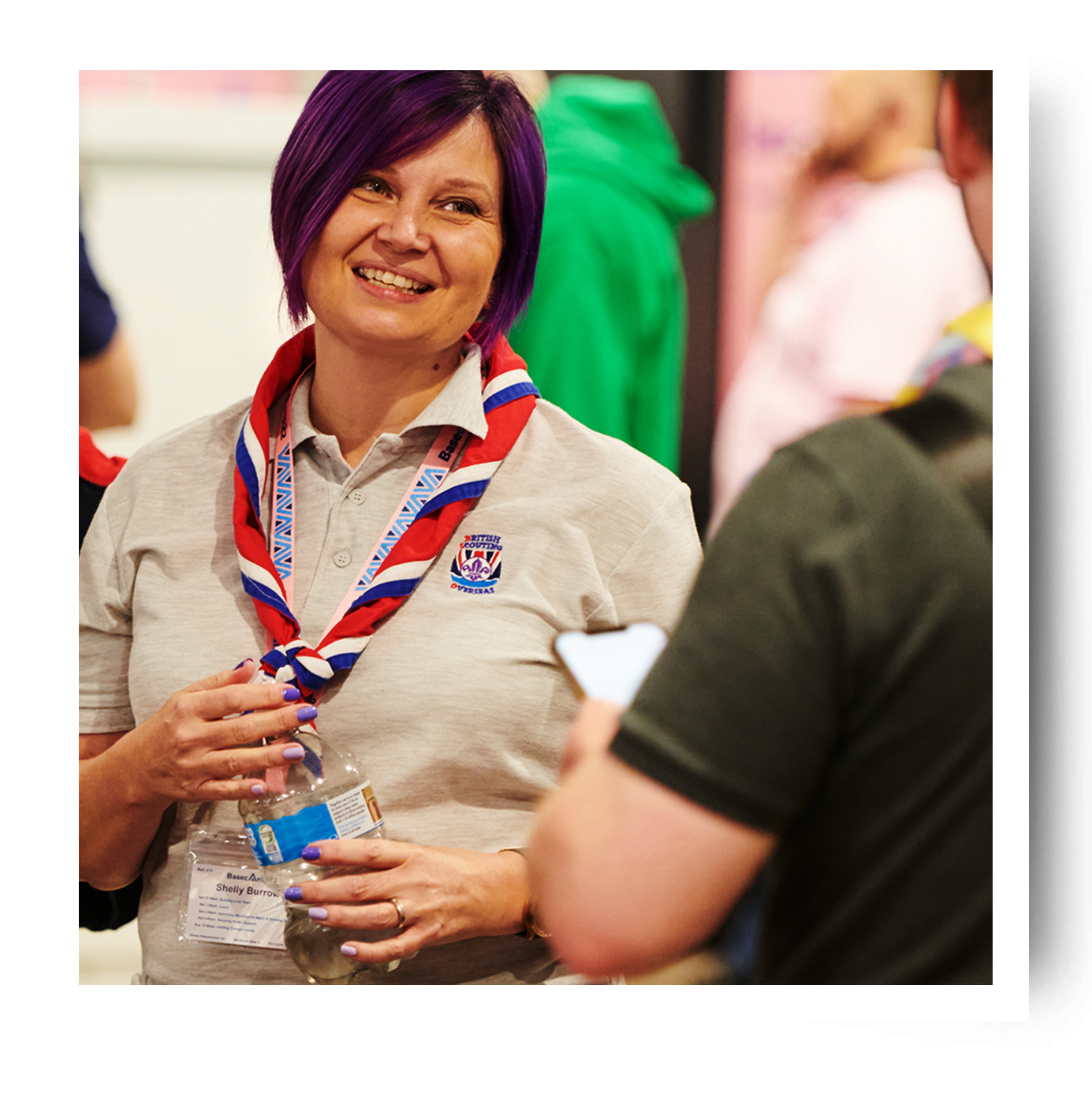 A volunteer with short purple hair is looking at another volunteer and smiling. They're wearing their uniform, a lanyard, and a blue, red and white necker.