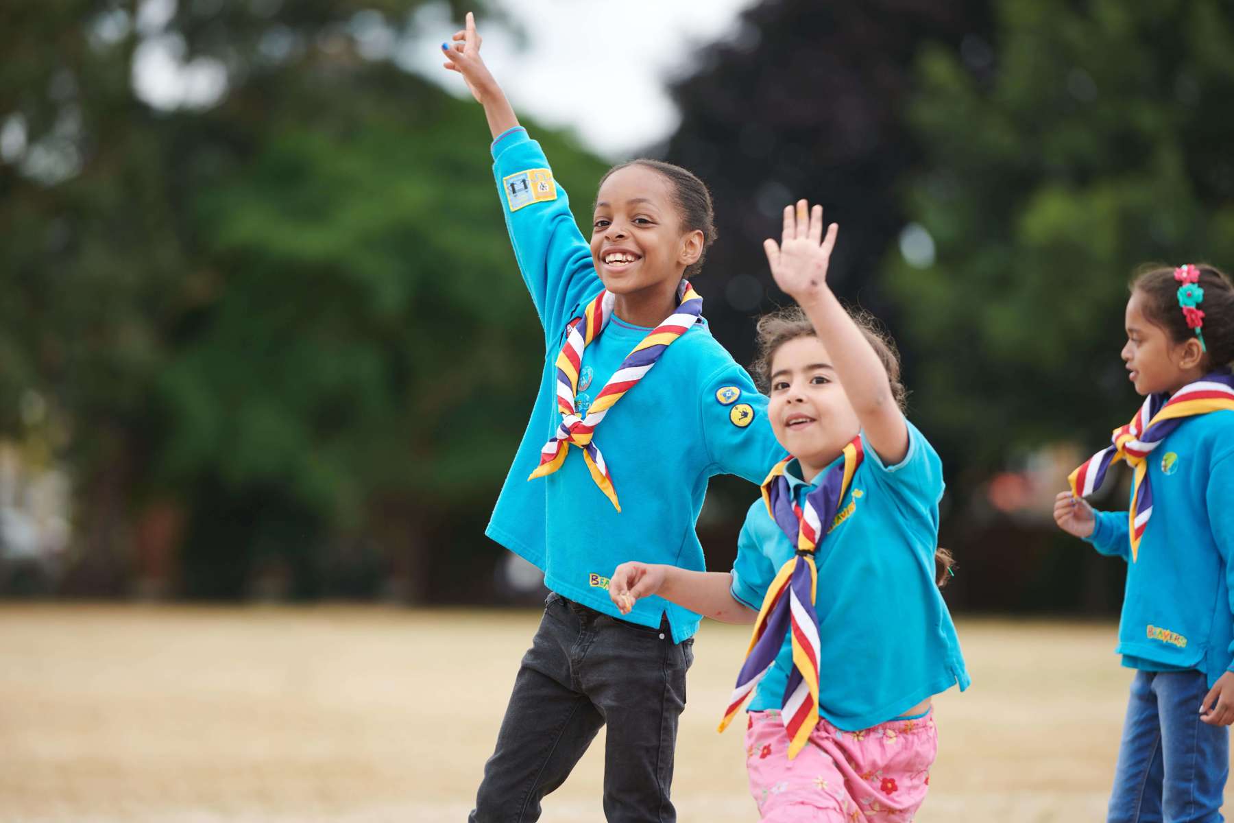 Two girl Beavers have their hands in the air, on a field outside their meeting place. They are smiling, with trees in the background.