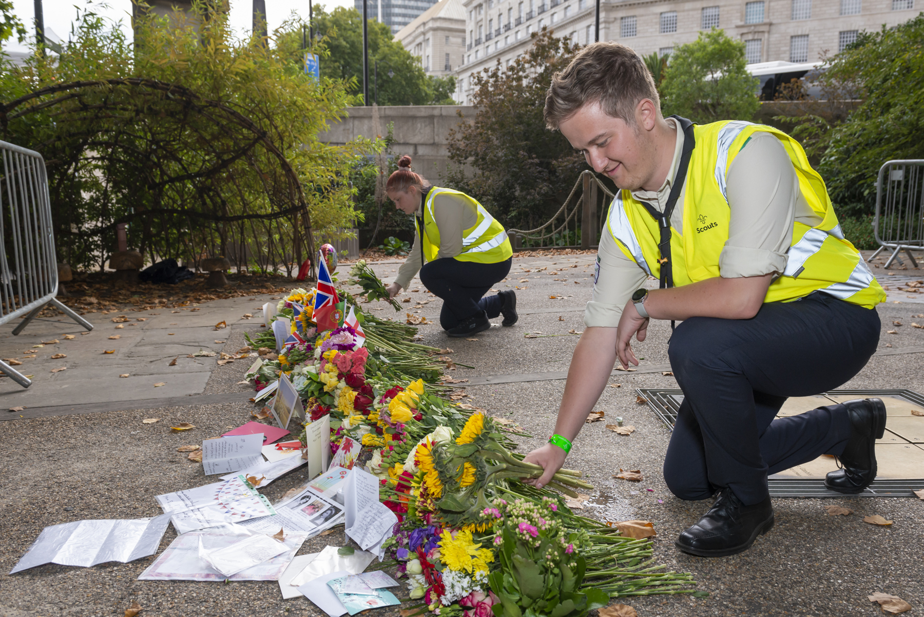 Two Scouts volunteers in hi-vis jackets are laying out flowers and cards in a row on the ground