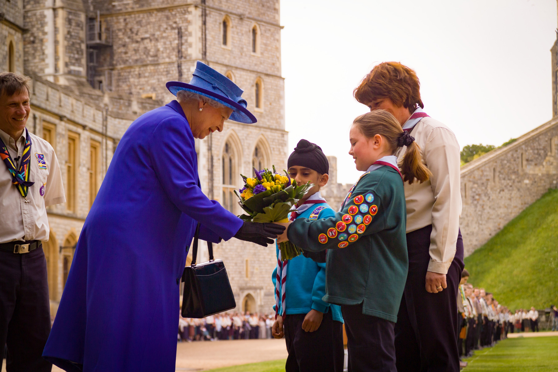 A Scout and Beaver hand over yellow and purple flowers to HM The Queen outside Windsor Castle