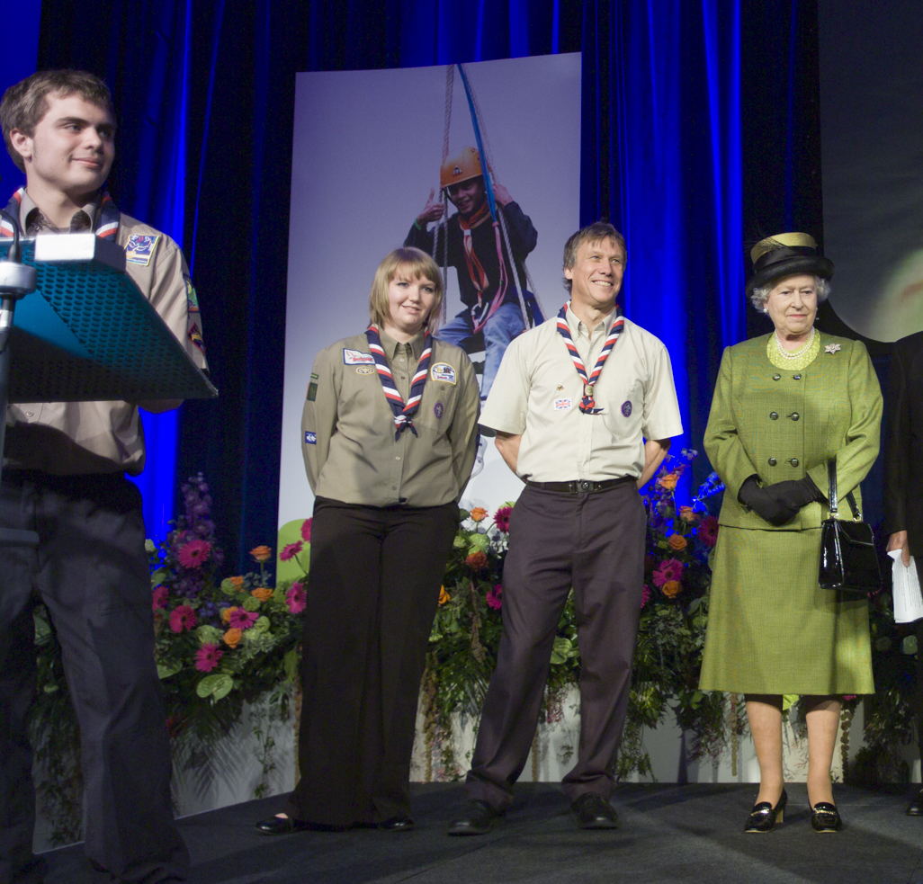 Amy, an Explorer Scout, stands with former Chief Scout Peter Duncan with HM The Queen on a stage in front of flowers, smiling.