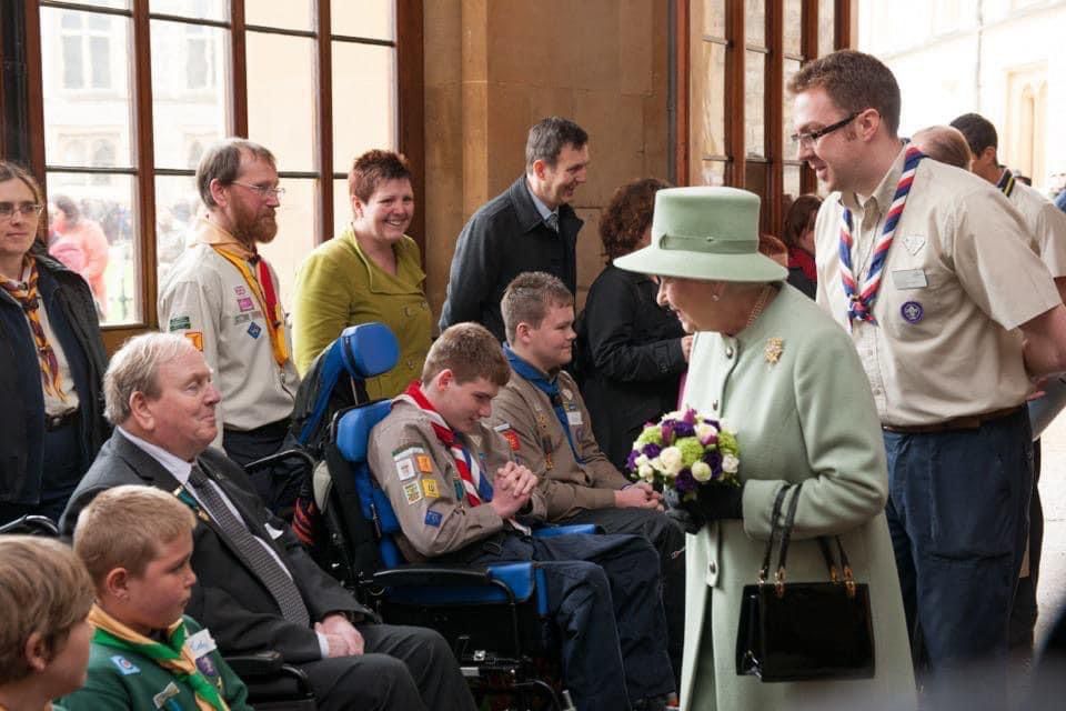 HM The Queen smiles at Scouts with flowers in her hand at Windsor Castle, with Scout volunteer Matt Rooney stood behind her.