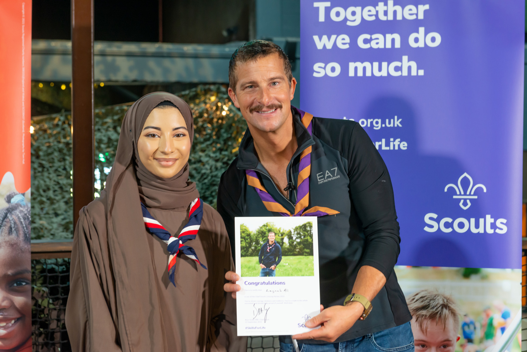 Chief Scout Bear Grylls stands with Scout volunteer Zaynab. Both are smiling at the camera and wearing neckers, and Bear holds a certificate saying 'congratulations.'