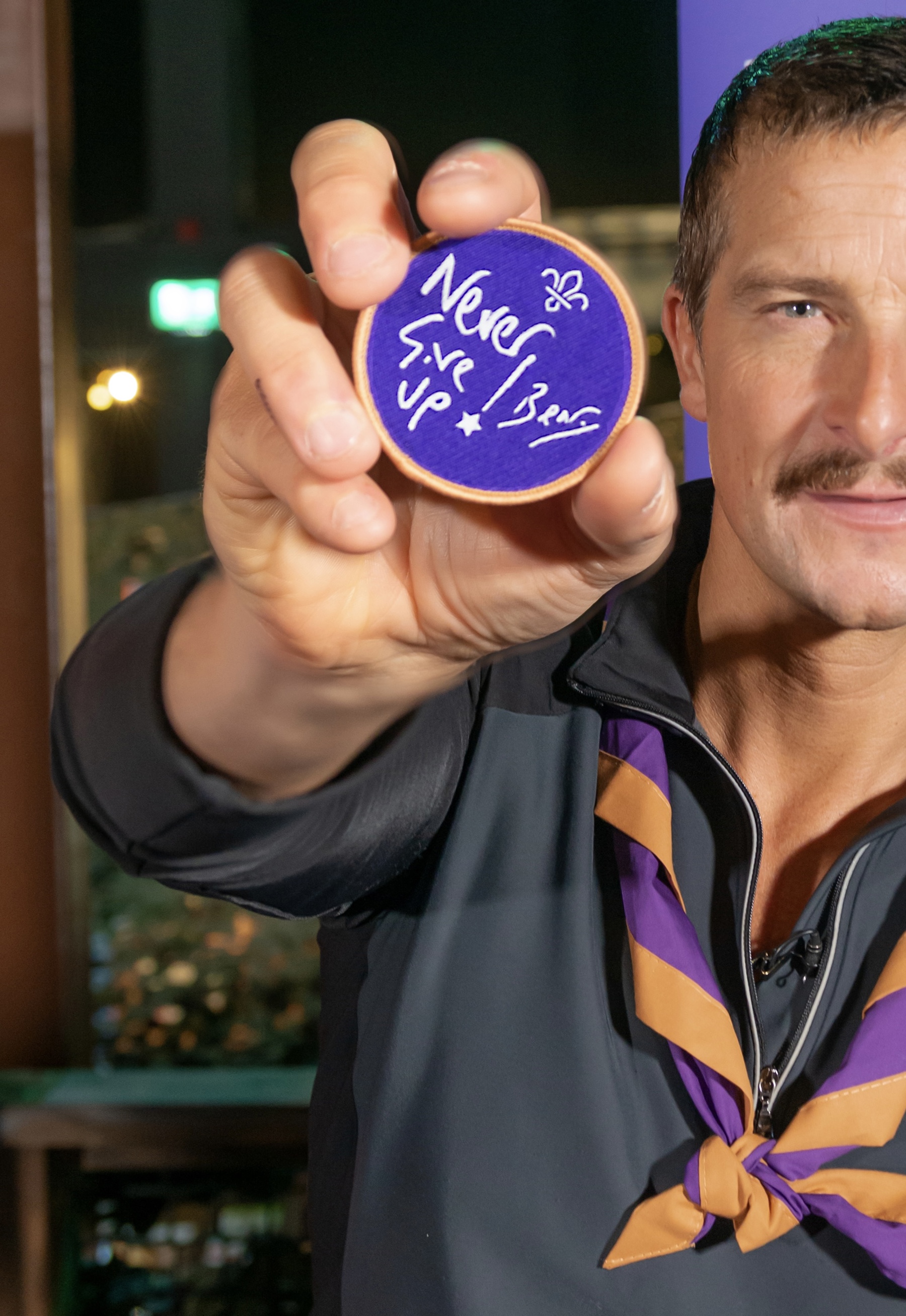 Bear Grylls holds up the Never Give Up cloth badge towards the camera