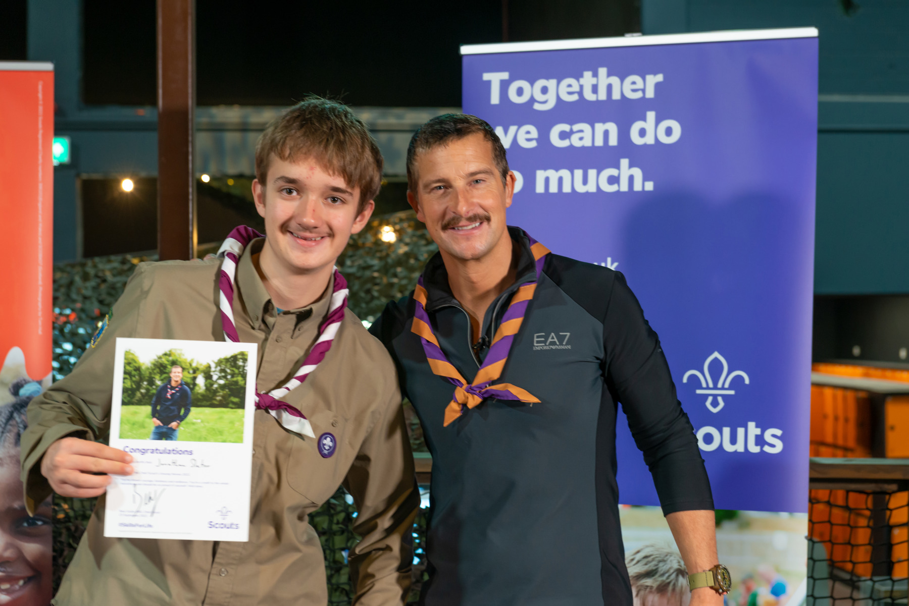 Chief Scout Bear Grylls stands with Scout Jonathan. They're both smiling at the camera and Jonathan is holding his congratulation certificate.