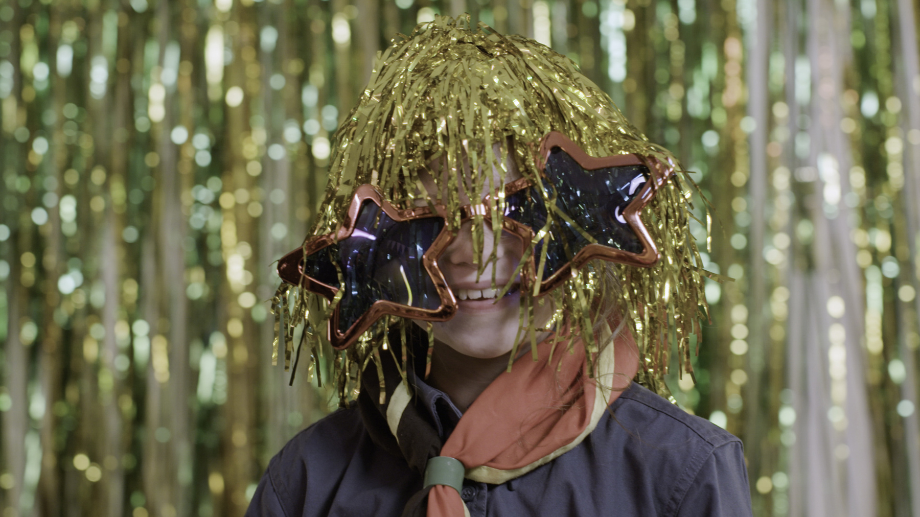 A young Squirrel is smiling while wearing a sparkly, gold tinsel wig and big, red, star shaped glasses. He is wearing a necker and against a gold tinsel backdrop.