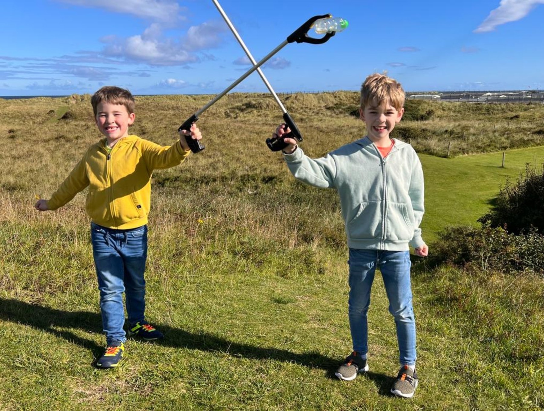 A Cub and Beaver are stood on grass with litter pickers in their hands as they pick up rubbish in their local area. Both are wearing jeans and jackets and smiling at the camera.