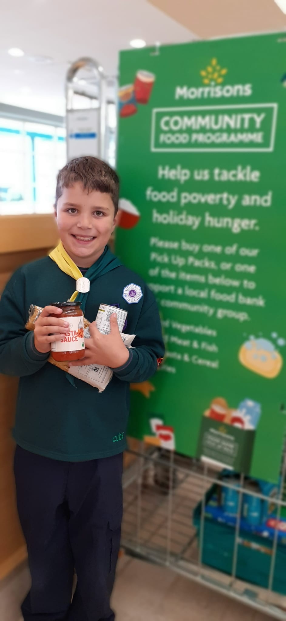 A Cub in uniform smiles as he stands by a food bank in his local supermarket, holding different items such as pasta and pasta sauce.