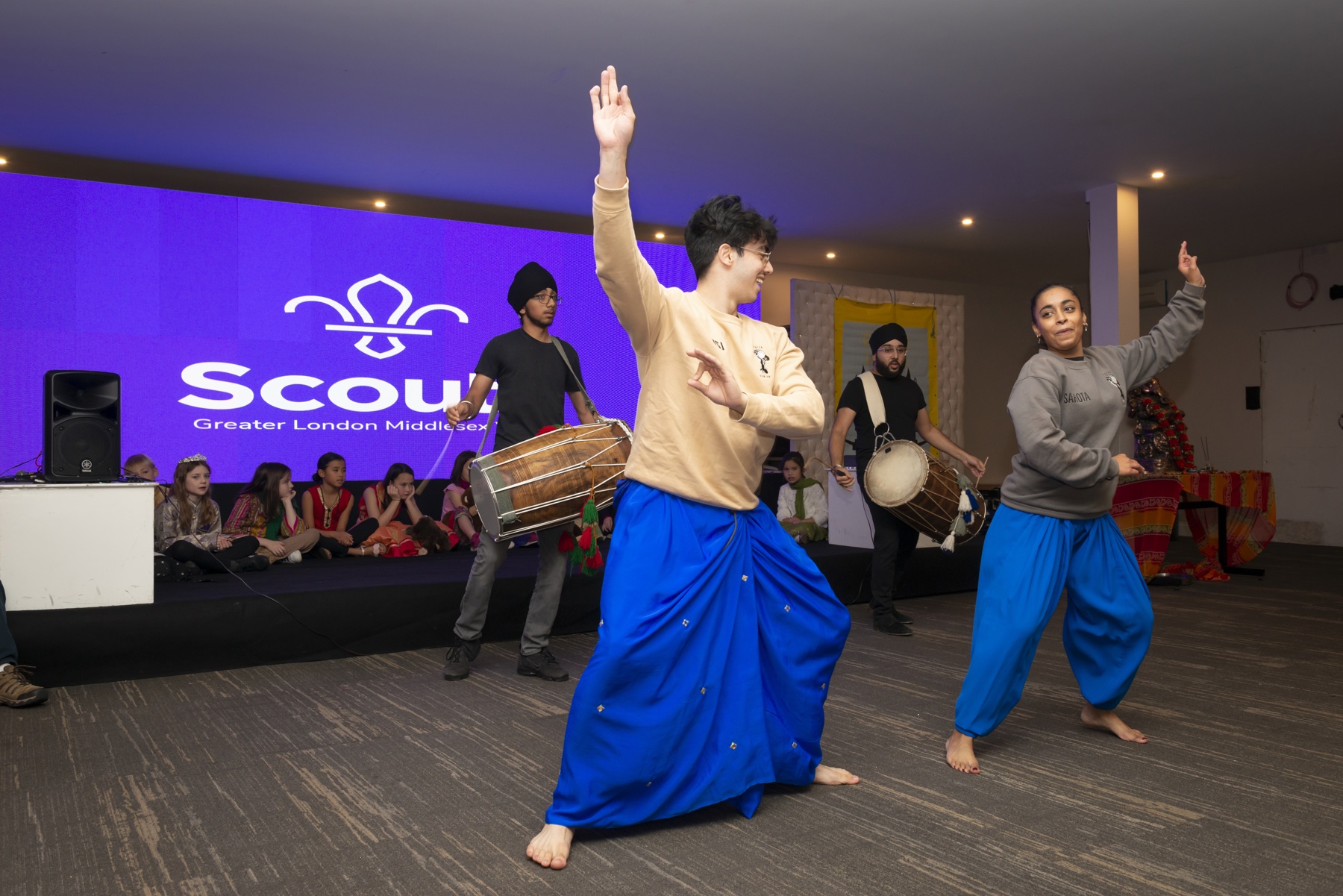 Two Scouts perform Bhangra dancing, in front of two other Scouts drumming. The dancers look at each other with their arms reaching out, wearing traditional Diwali clothing