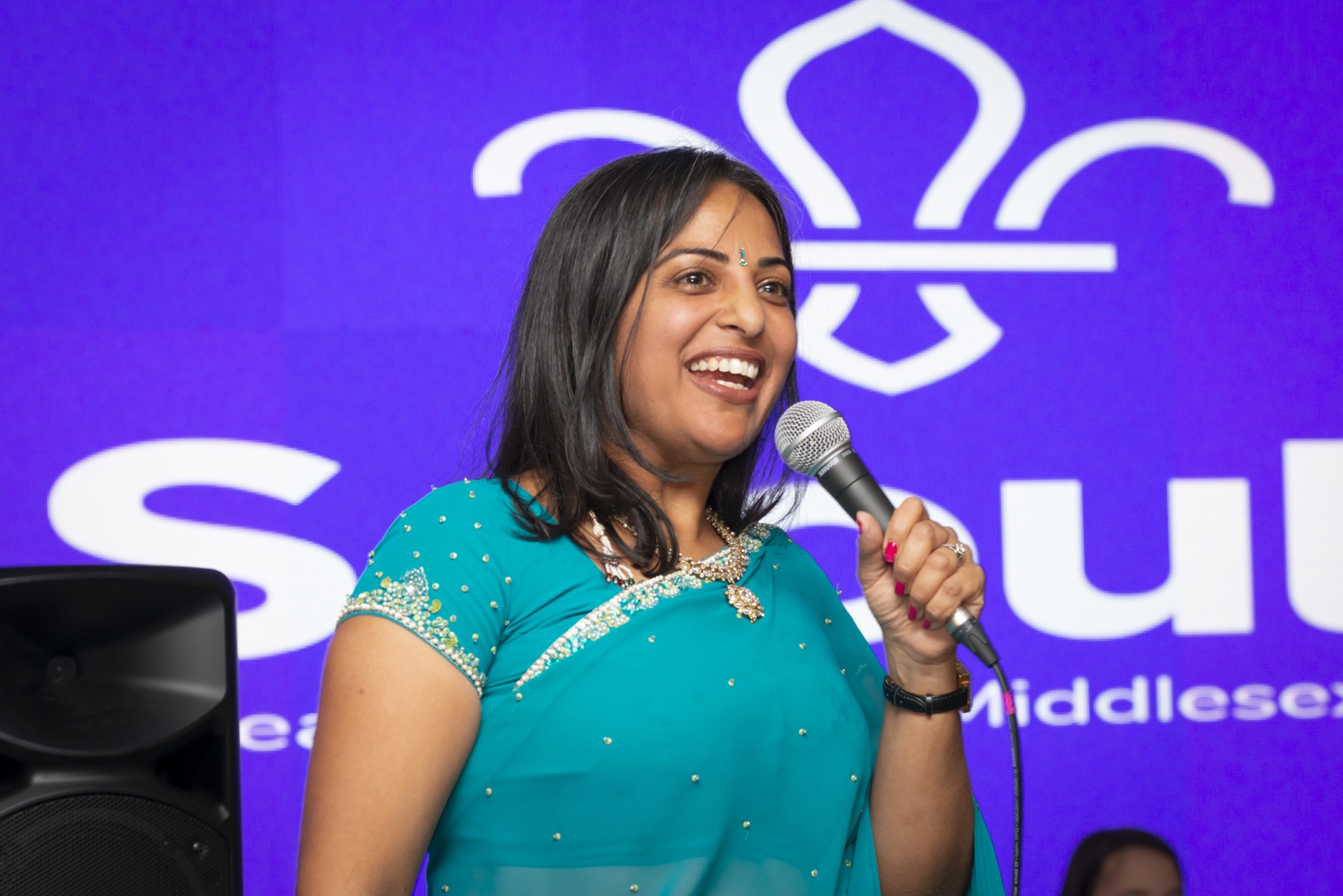 UK Commissioner for Perception, Nisha Patel, smiles while holding a microphone and wearing traditional Diwali clothing