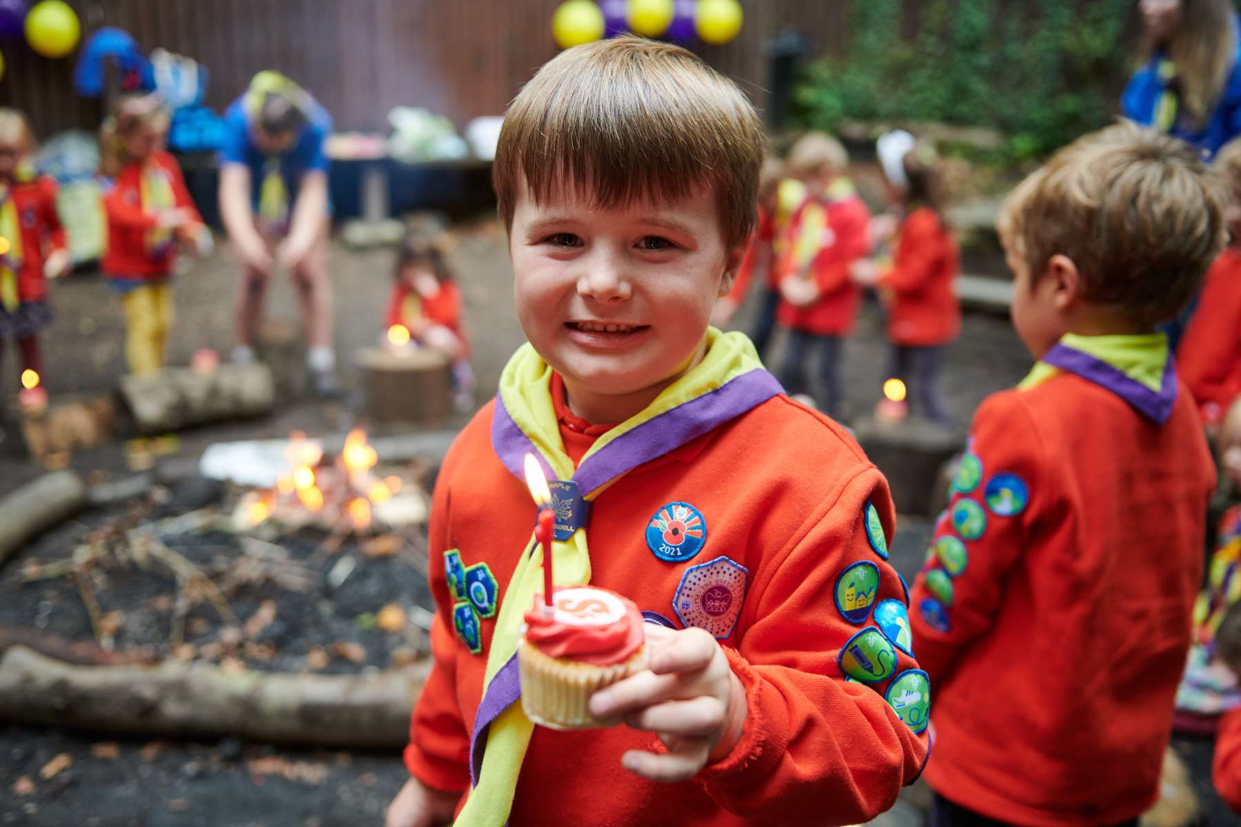 A Squirrel wearing a necker and uniform is holding a cupcake with the Squirrels logo on and a candle in, with other Squirrels and a bonfire in the background