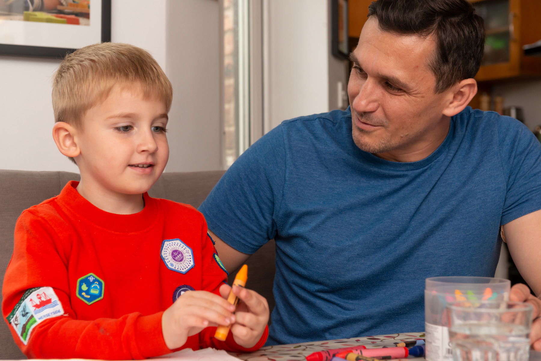 Oscar is holding an orange crayon and wearing his Squirrel jumper and looking at a box of crayons. He's sitting next to his dad who's looking at him, wearing a blue t-shirt.