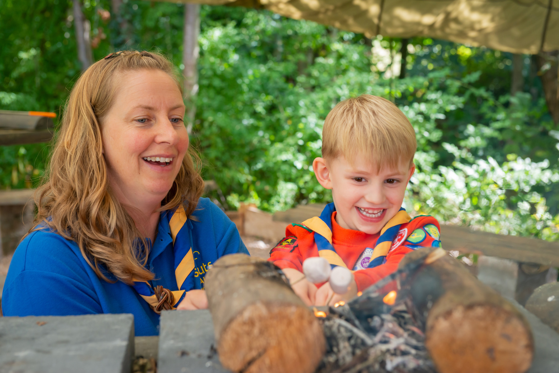 Louise, a Squirrels volunteer, is wearing a blue polo shirt with a necker while helping her son, George, in Squirrels uniform, toast marshmallows. They're both looking at the marshmallows and smiling.