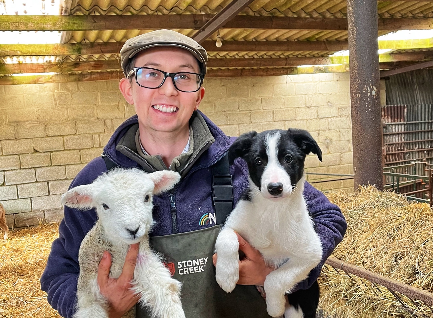 Karl Franklin, a farmer, is holding a lamb in his right hand and a black and white sheep dog in his left hand, and is looking at the camera smiling. He's wearing a flat cap and glasses and is stood in a lambing shed filled with hay.