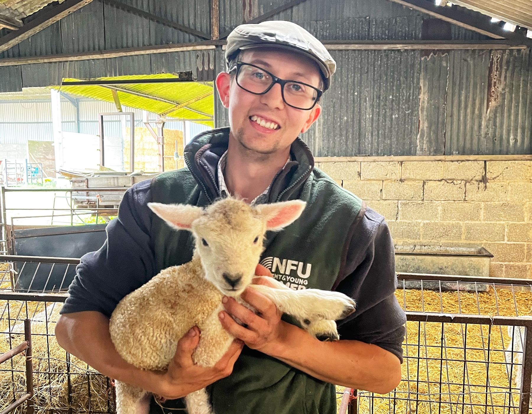 Karl Franklin is holding a lamb in his arms and is smiling at the camera. He's stood in a lambing shed filled with hay and is wearing glasses and a flat cap.