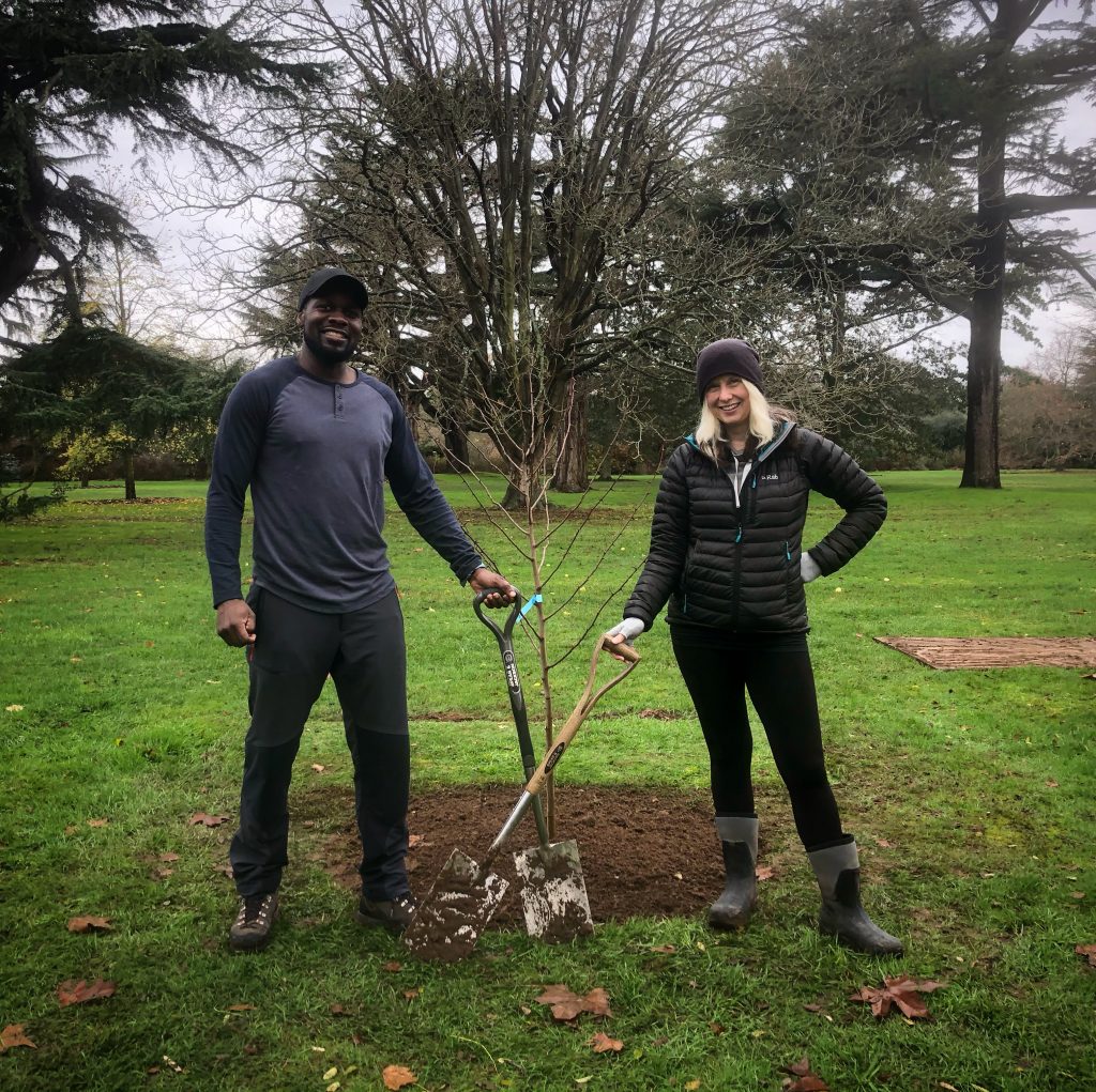 Dwayne Fields and Phoebe Smith are standing on grass holding shovels after planting a tree together at Kew Gardens.