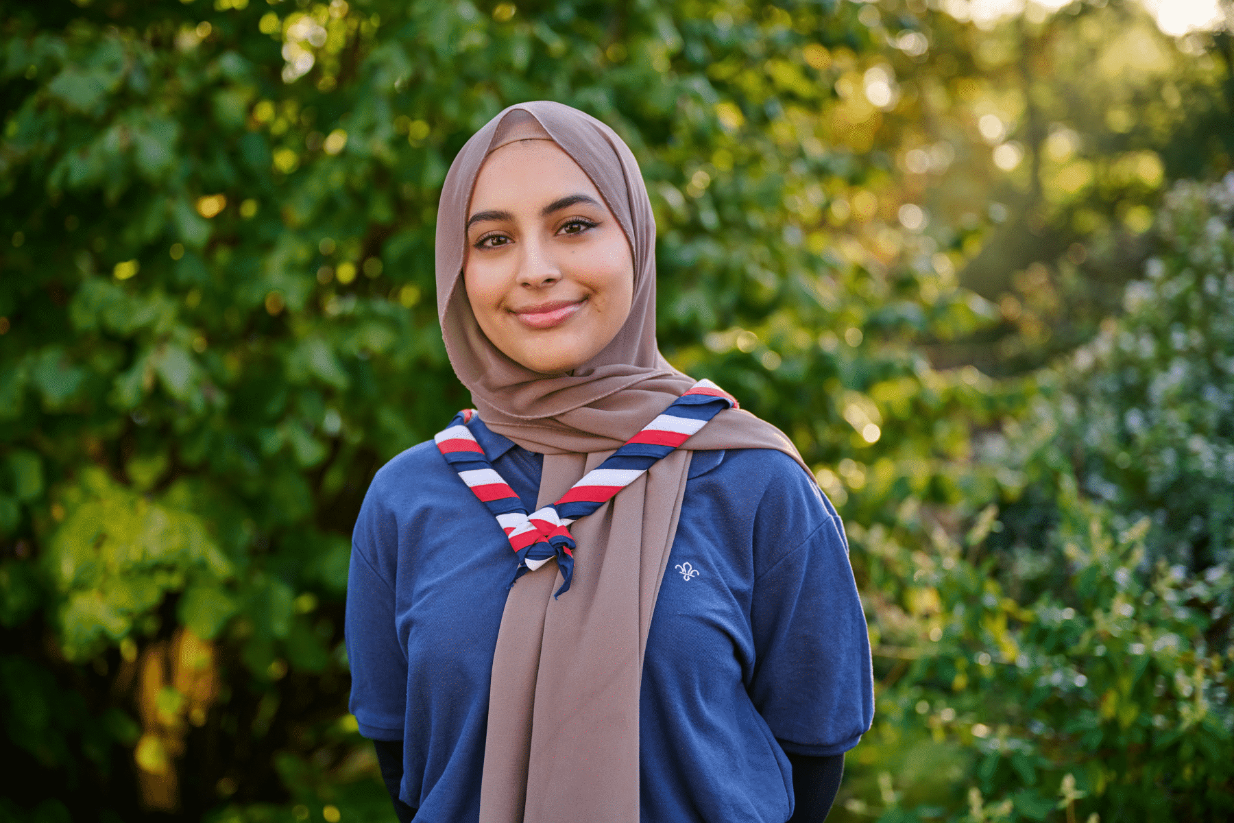 Ayesha is stood in front of trees. She is smiling and is wearing her hijab and Scouts necker. She looks relaxed and is wearing a blue Scouts top, with her hands behind her back.