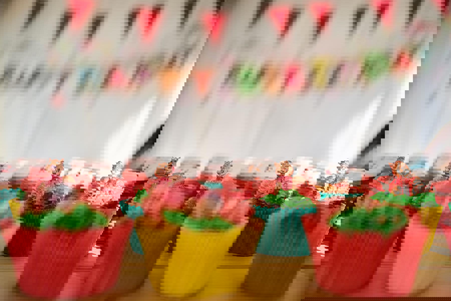 There are cupcakes are on a table, with a happy birthday banner in the background. The cakes have yellow, blue and red wrappers, with green icing and Squirrel decorations.
