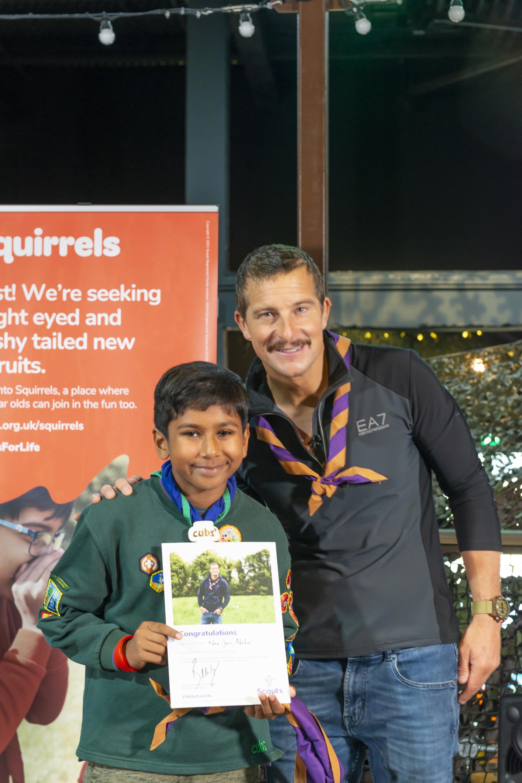 Neo is stood next to Bear Grylls and holding his Unsung Heroes certificate. They're both smiling. Bear is wearing a necker. Neo is wearing his Cubs uniform.